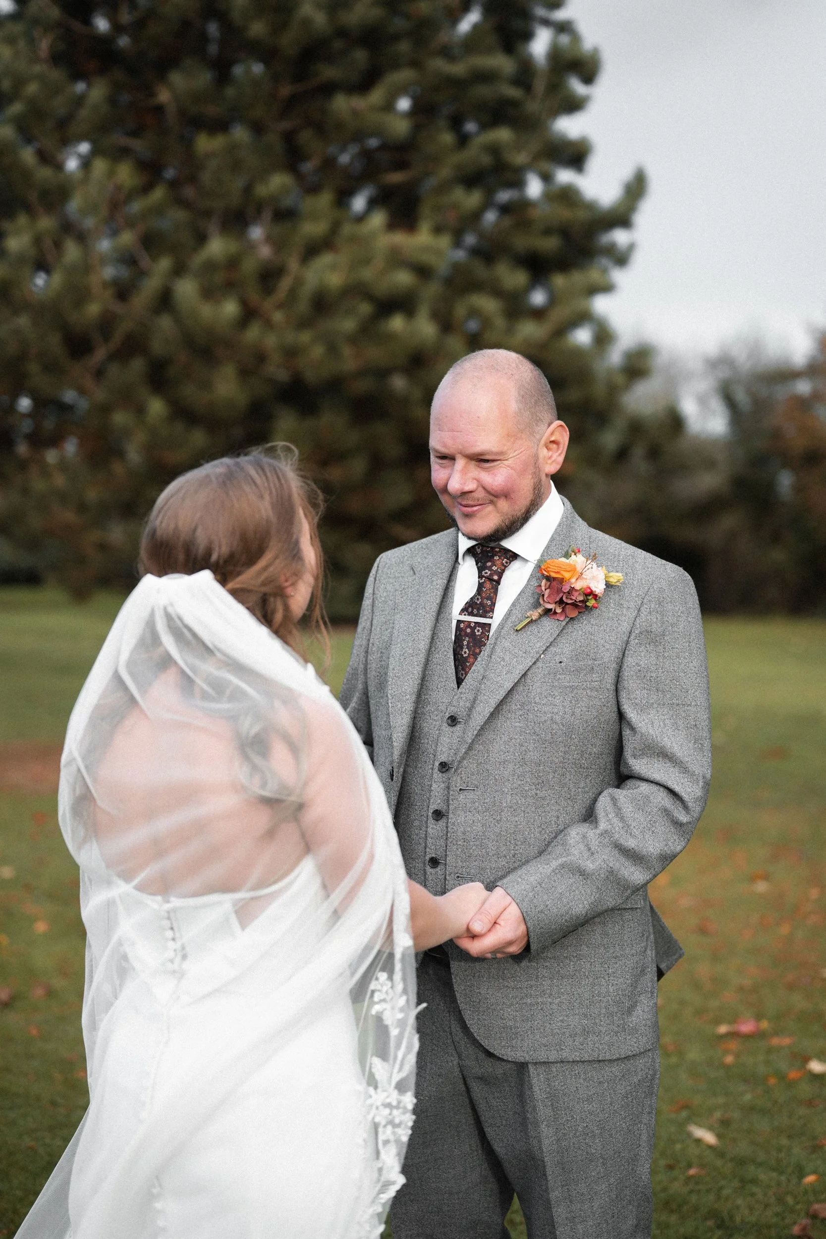A bride and groom holding hands during their outdoor wedding ceremony, with a large tree in the background.