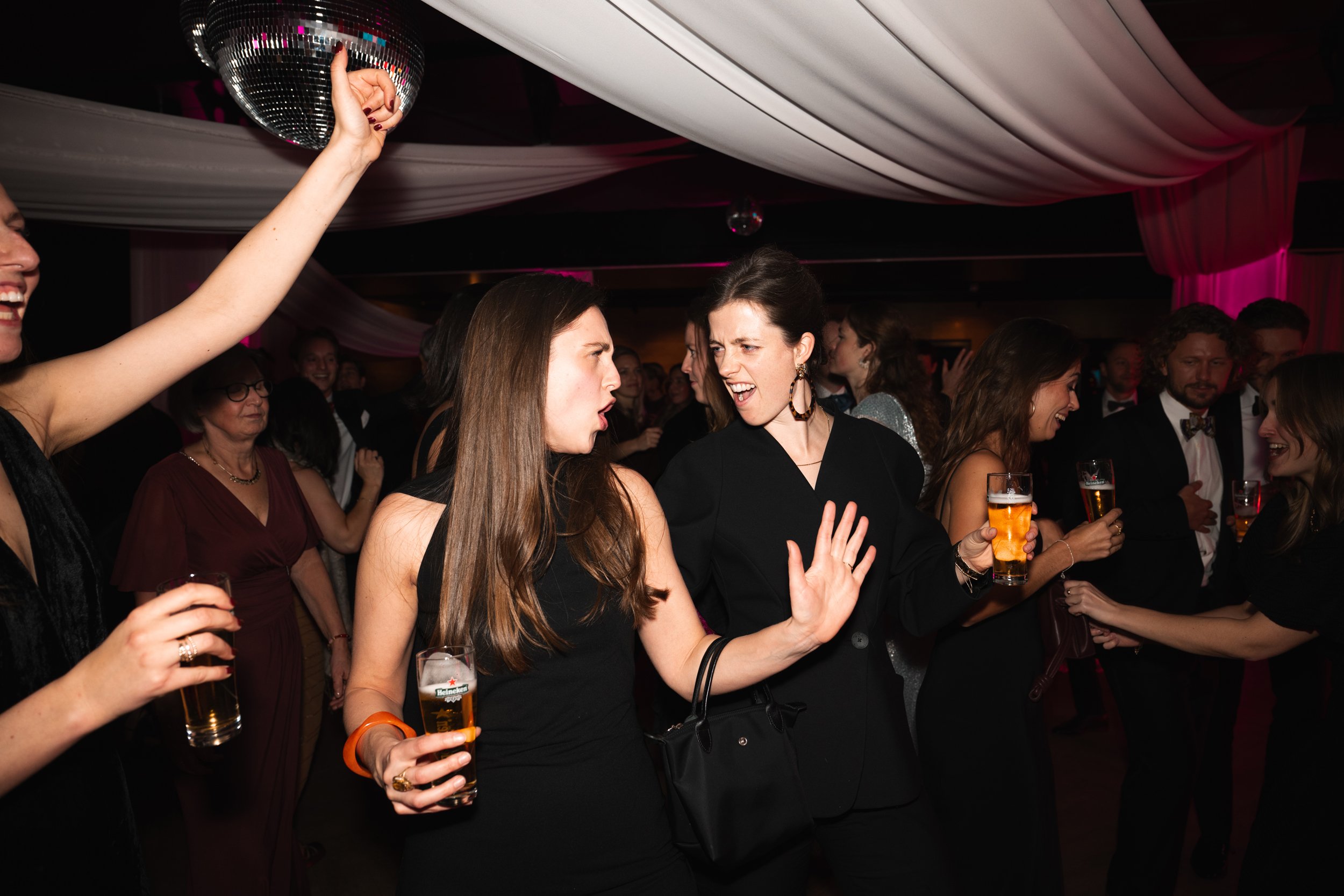 People dancing and socializing at a party with a disco ball, drinks in hand, under draped ceiling decorations.