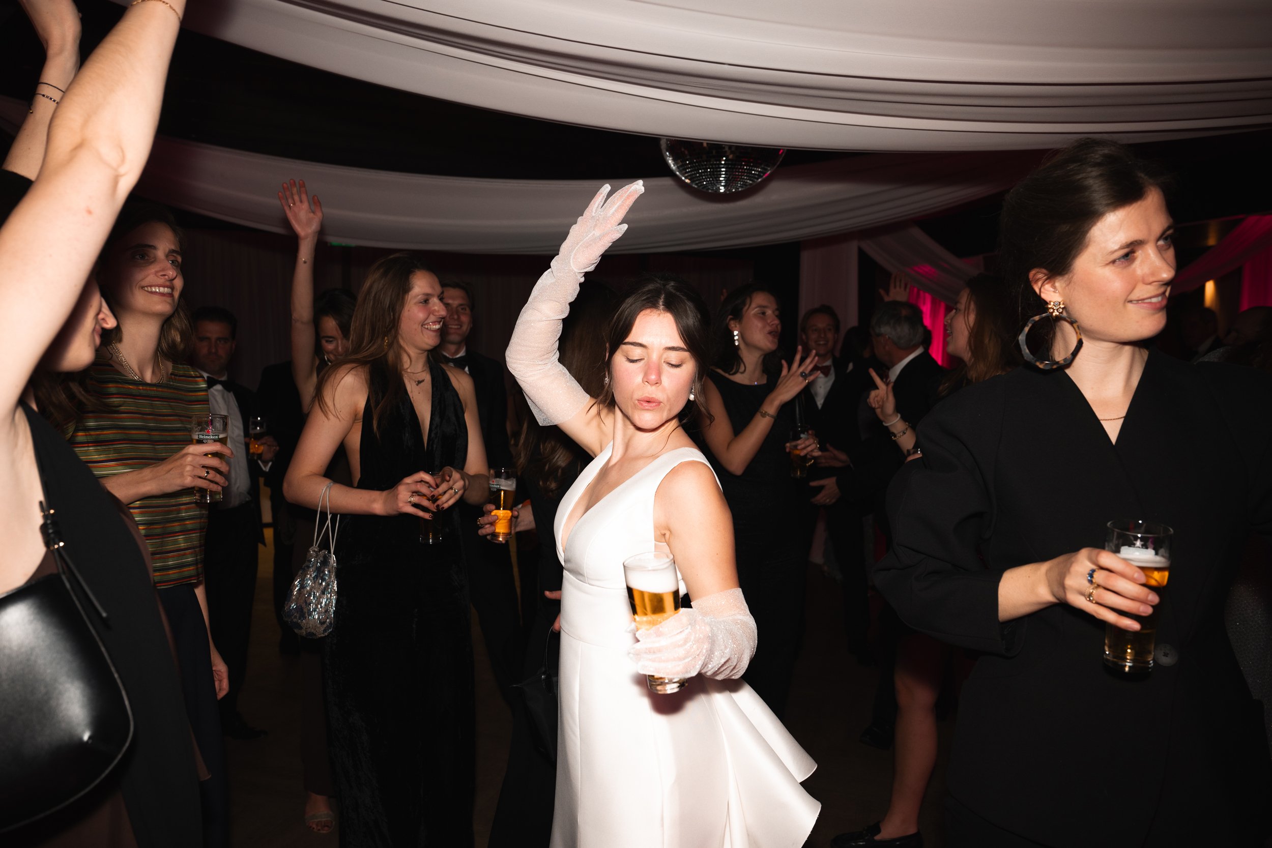 People dancing and socializing at a party or wedding reception, with a woman in a white dress in the center holding a glass of beer.