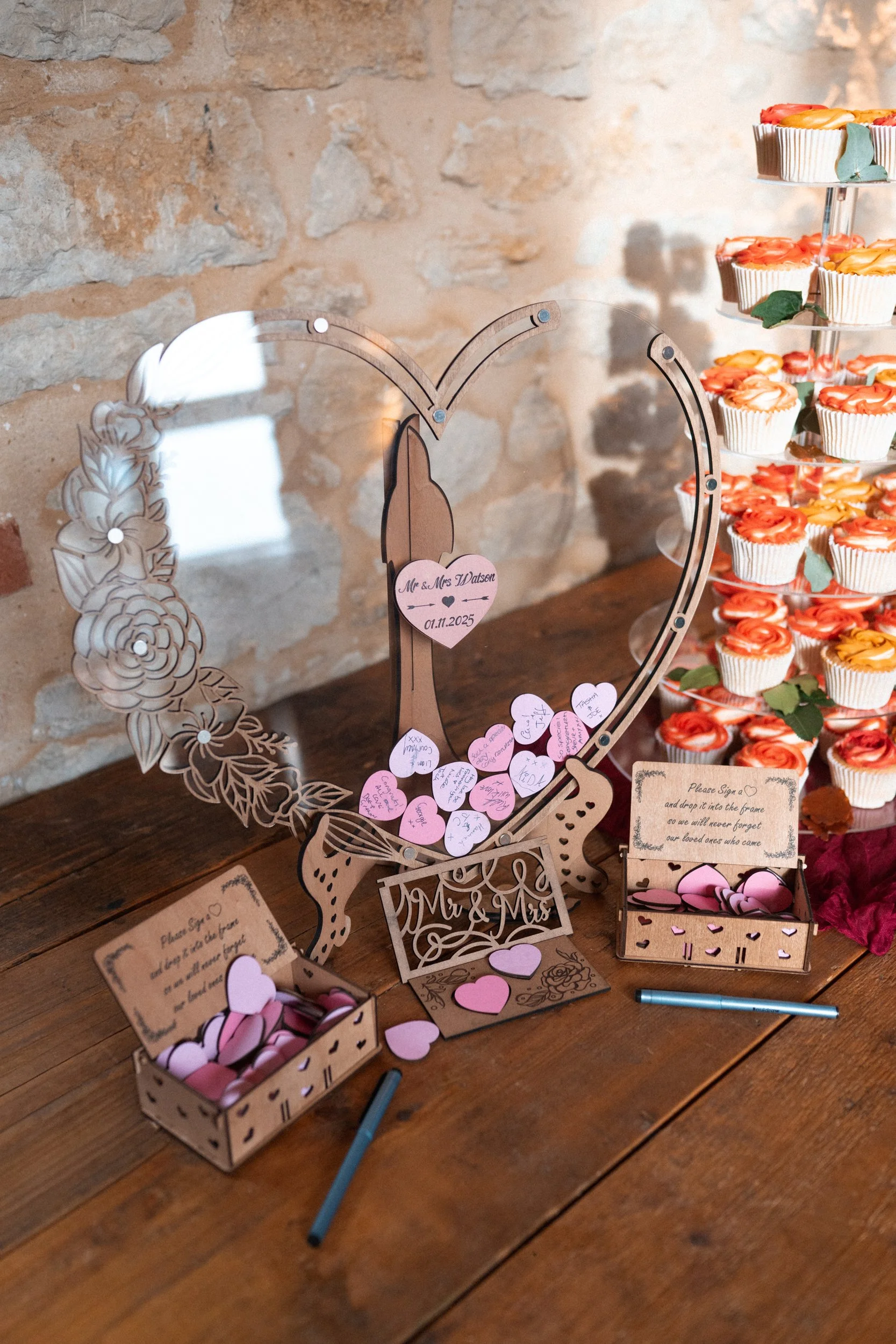 Wedding guestbook with pink hearts on a table, with a cupcake stand filled with orange roses cupcakes in the background.