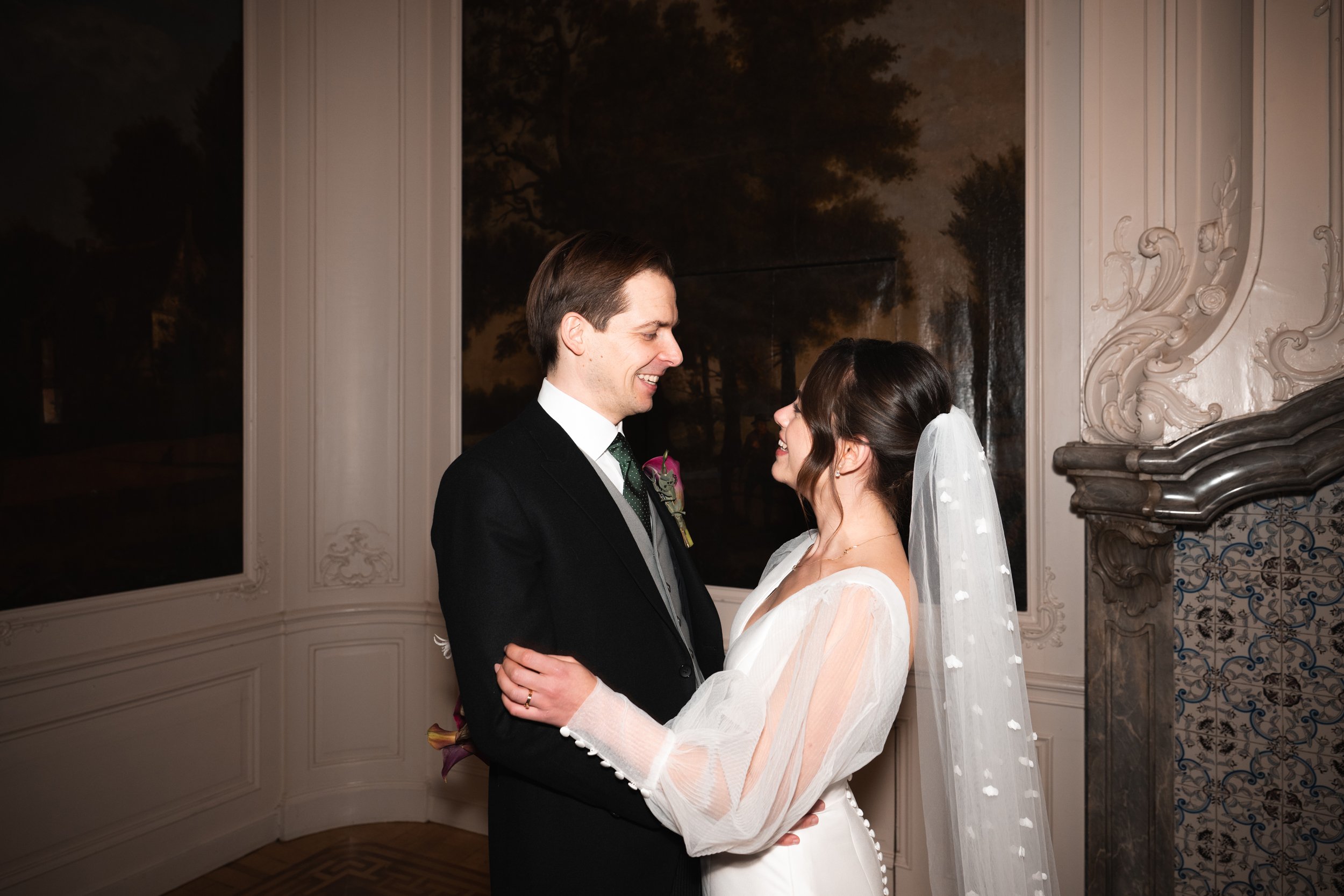 A bride and groom are holding each other and smiling during their wedding inside a decorated room with large windows and ornate wall details.
