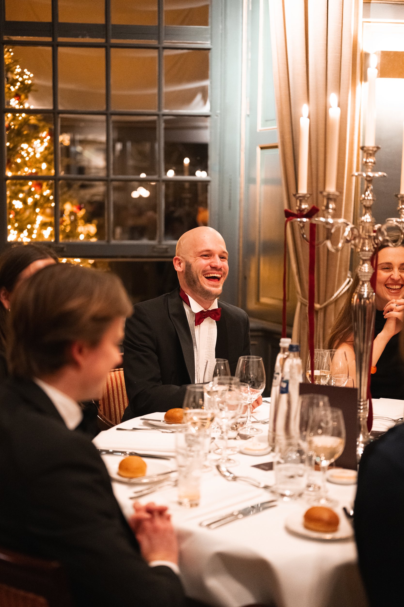 People dressed in tuxedos and dresses sitting at a dinner table celebrating with a Christmas tree visible through the window in the background.