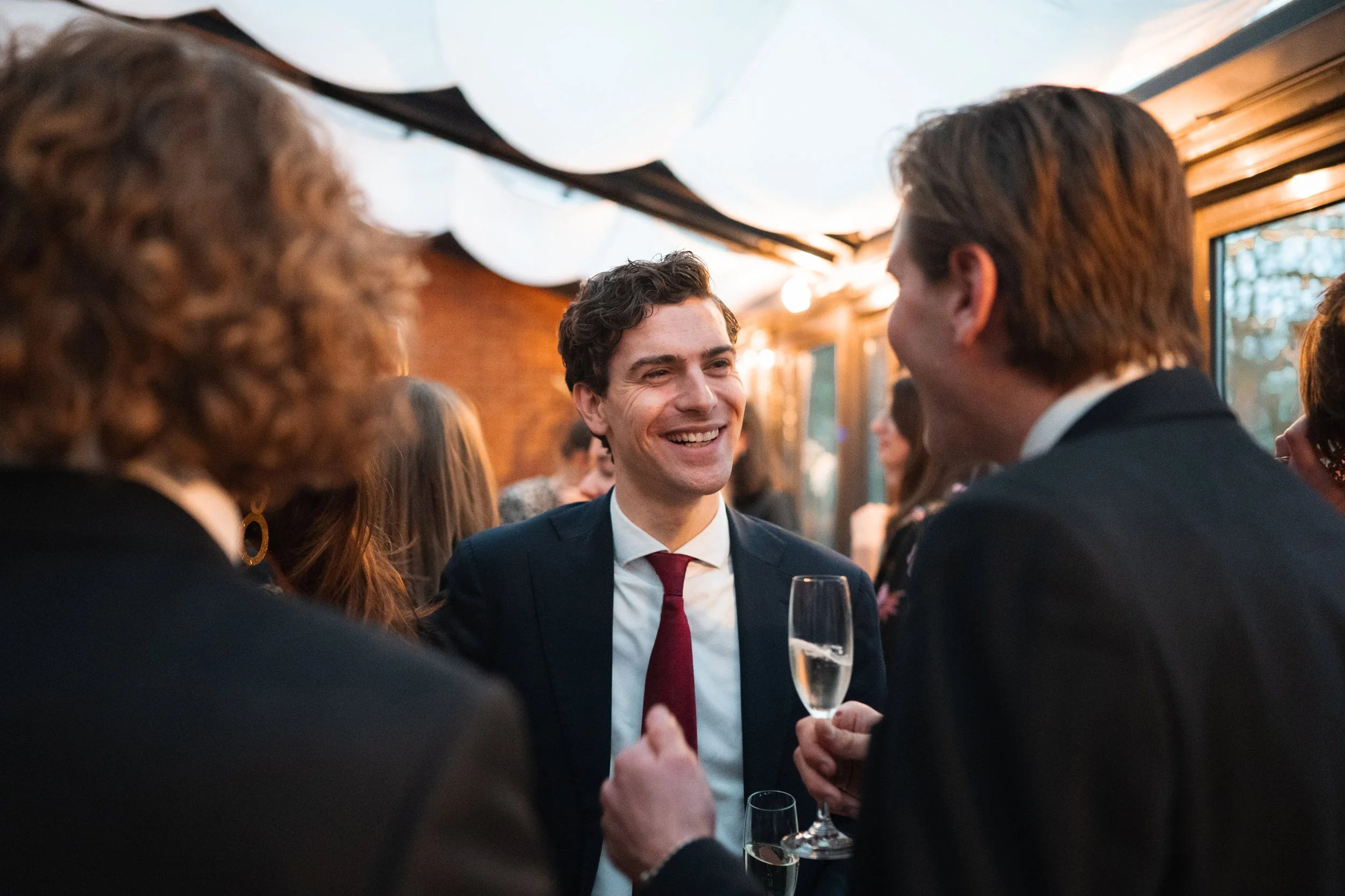 A man in a suit and red tie smiling and talking to a woman holding a champagne glass at a social gathering.