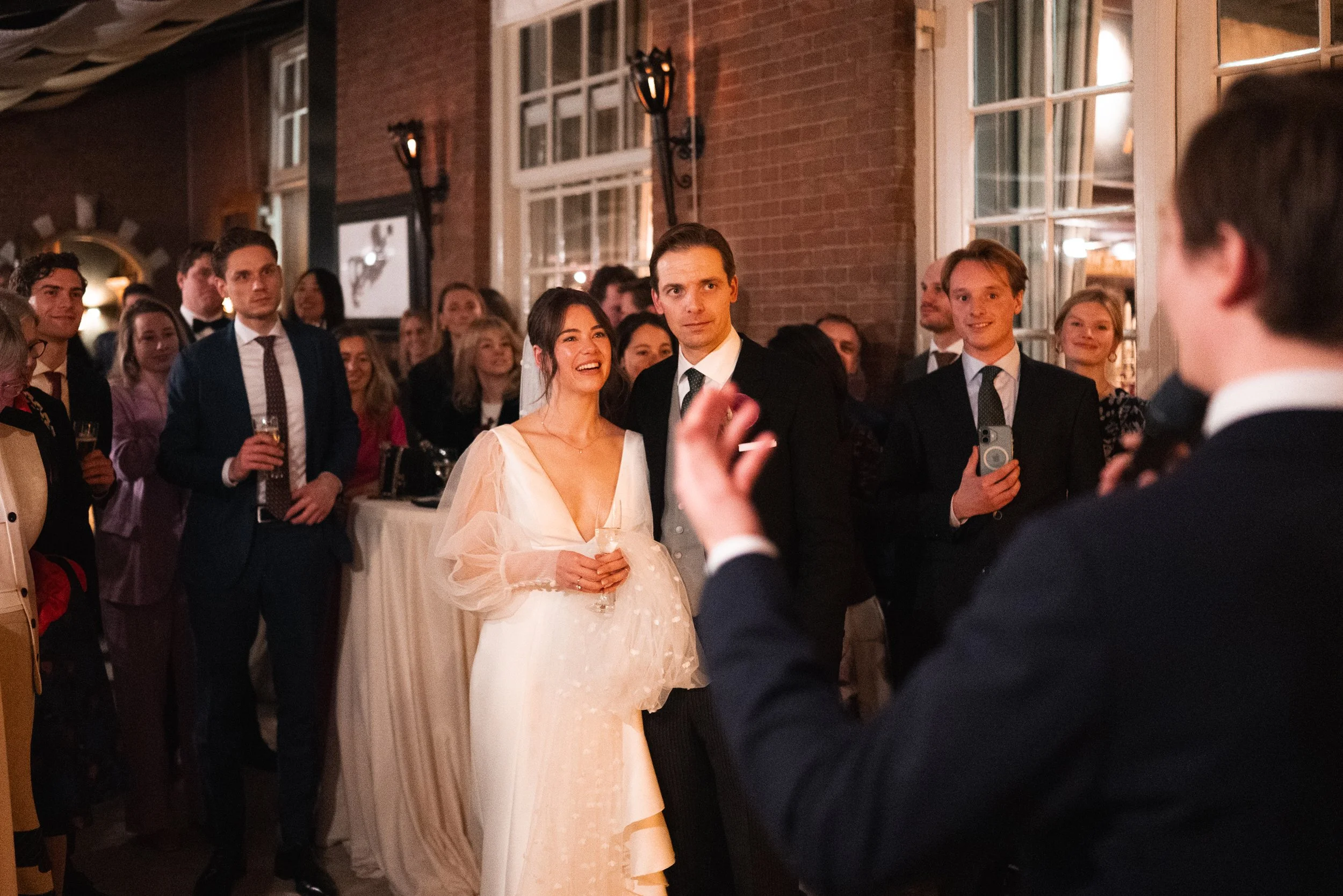 A bride and groom at their wedding reception surrounded by guests, with the groom speaking and the bride smiling, in a warmly lit indoor setting with brick walls and large windows.