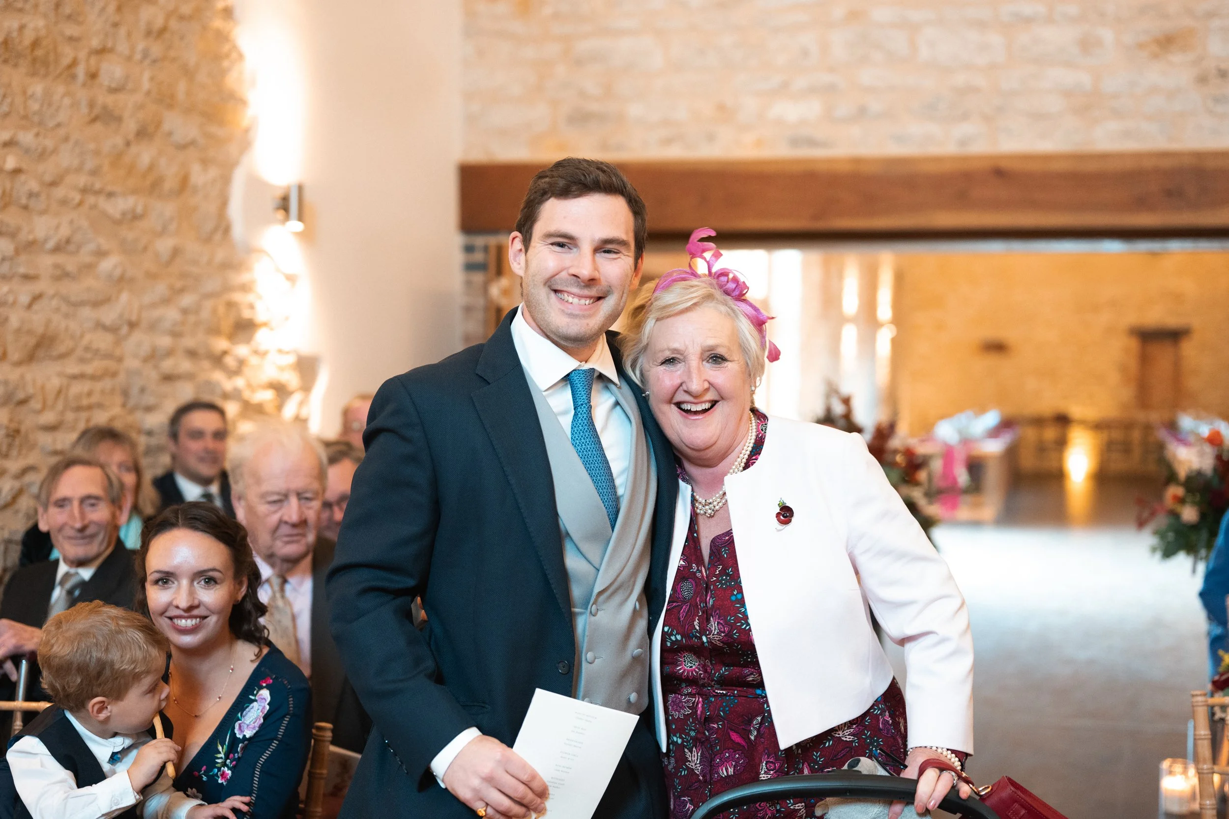 A young man and an elderly woman at a wedding or celebration, smiling and posing together indoors with a crowd of people in the background.