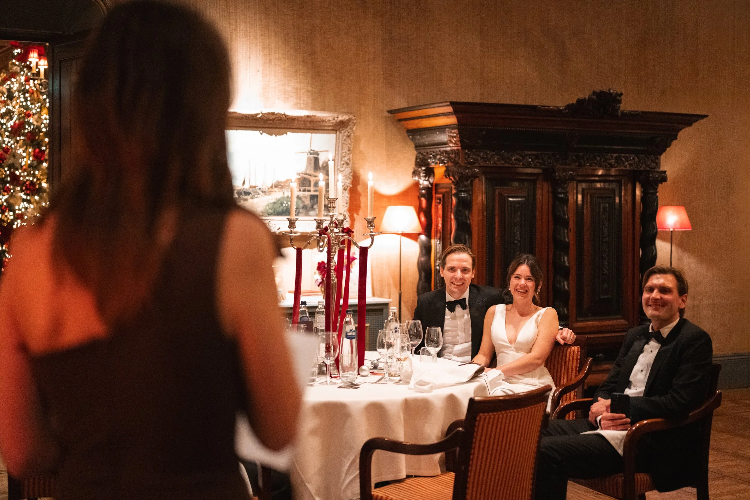 People sitting at a dining table during a formal event, with a woman in black dress in the foreground and three smiling individuals in tuxedos and a white dress in the background, in a decorated room with Christmas tree and antique furniture.