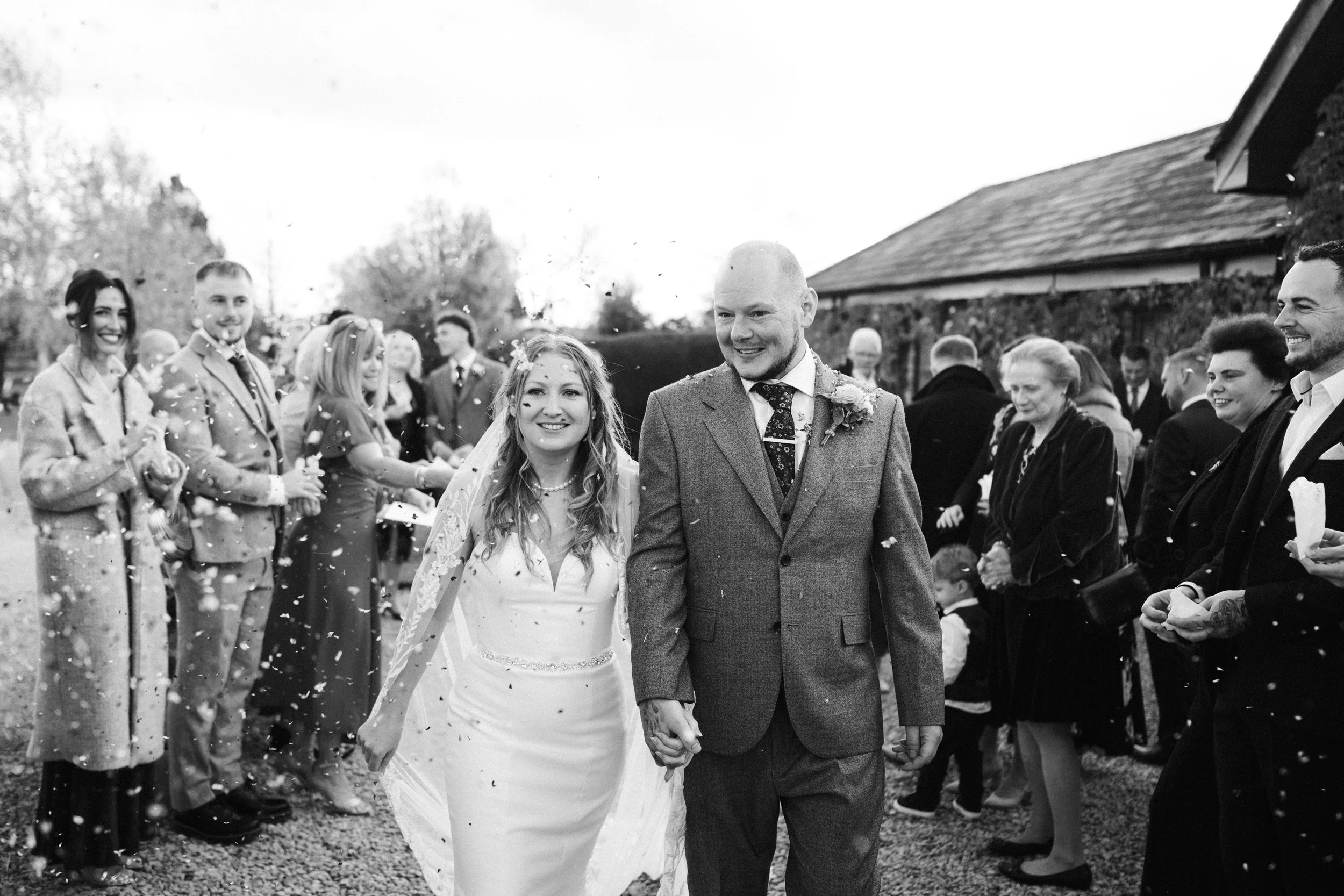 A black-and-white photo of a newlywed couple walking hand-in-hand outside, surrounded by smiling friends and family in formal attire, with confetti falling around them.