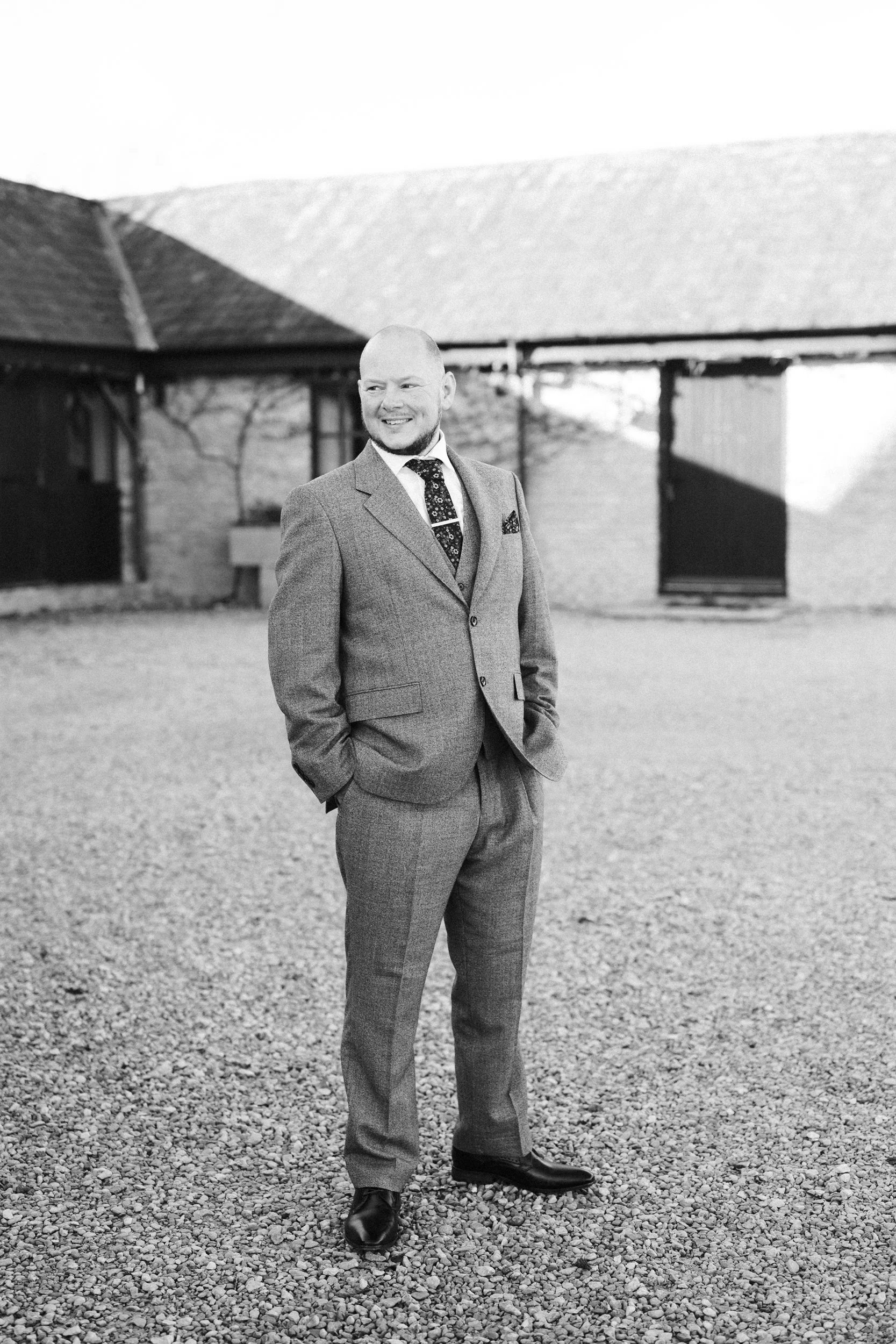 A man in a suit standing outdoors on a gravel ground, smiling, with a building in the background.