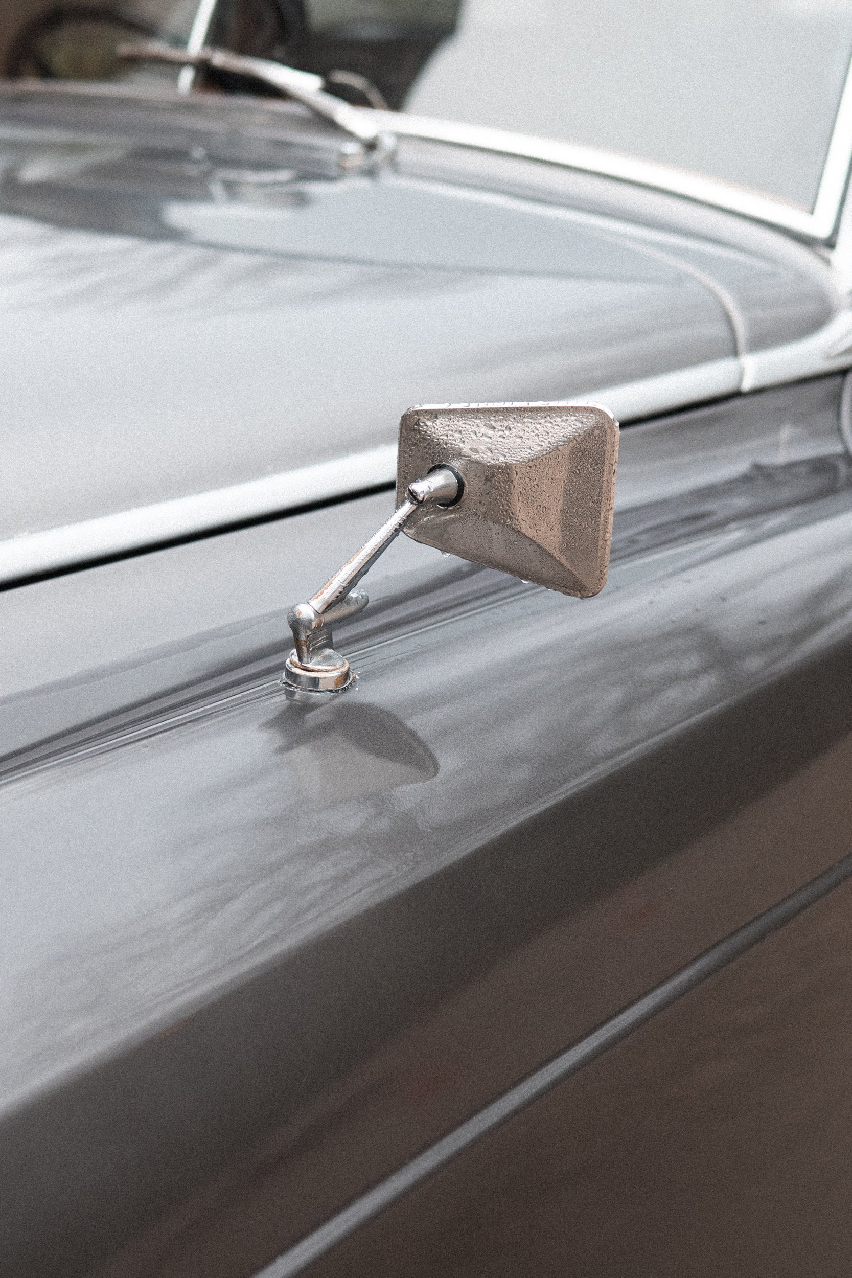 Close-up of a vintage car's side mirror mounted on the hood, with the front part of the car in the background.