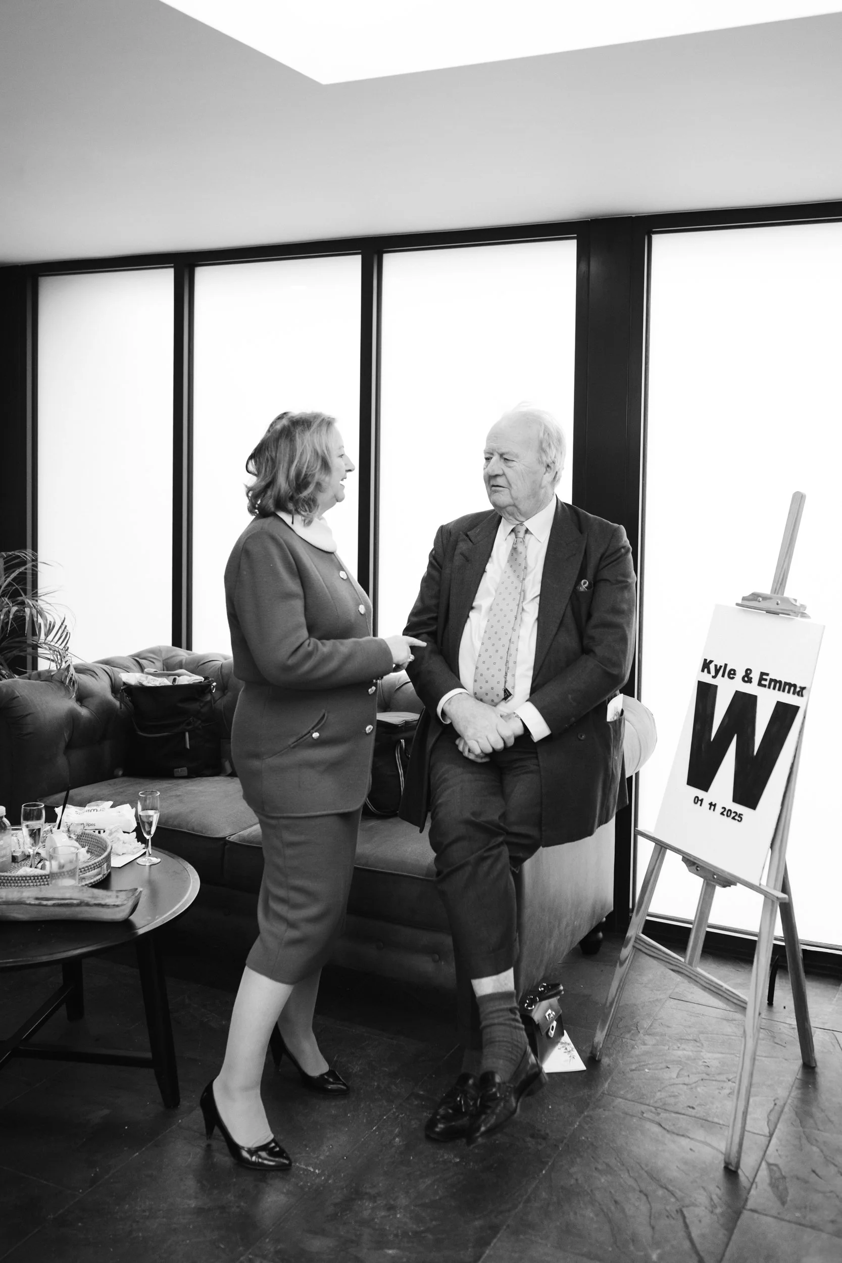 A woman and an elderly man converse in an indoor setting with large frosted windows. The woman is standing, wearing a tailored suit, and the man is sitting on a cushioned bench, dressed in a suit with a tie. There is a table with drinks and snacks ne
