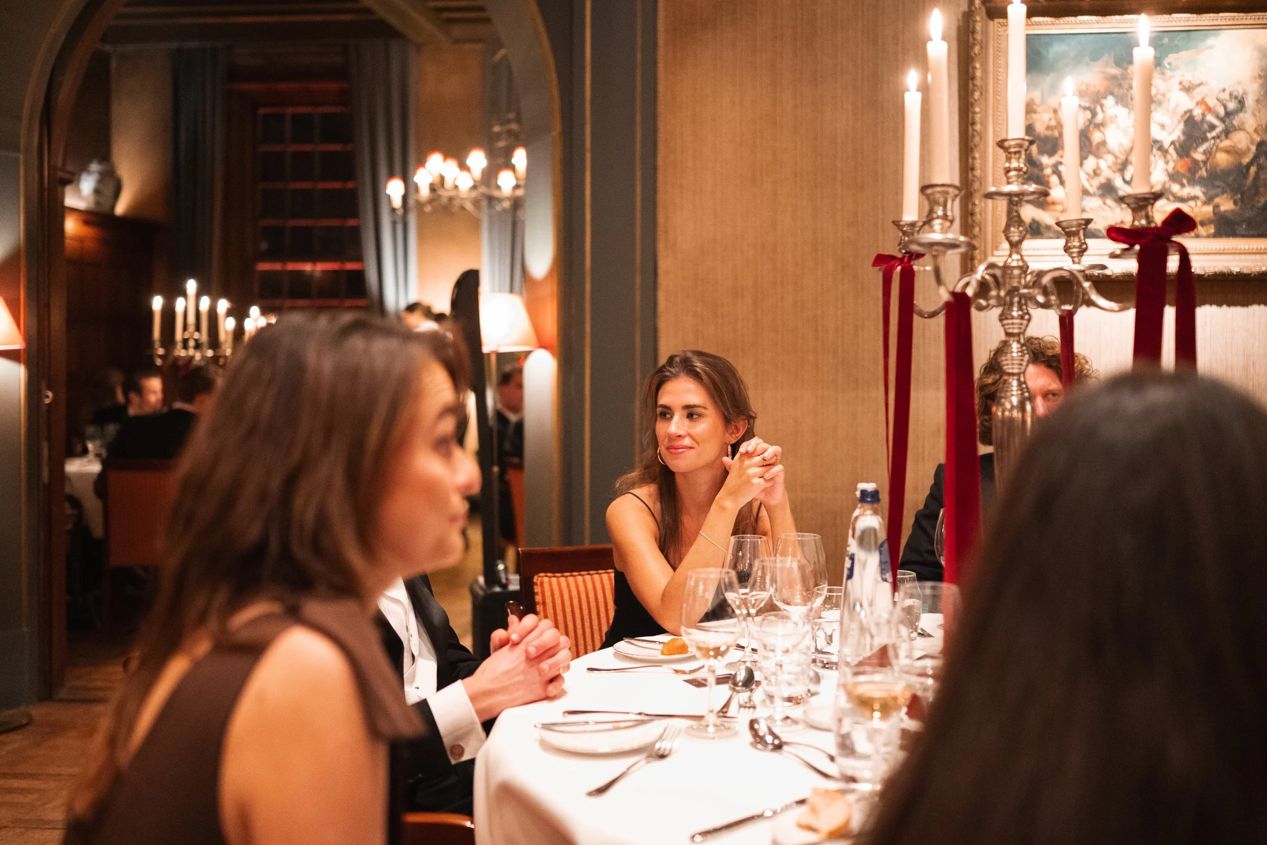 A woman with brown hair in a sleeveless dress sits at a dinner table with crystal glasses, surrounded by other guests in a warmly lit, elegant restaurant. A tall candelabrum with lit candles and red ribbons is on the table. The restaurant has artwork