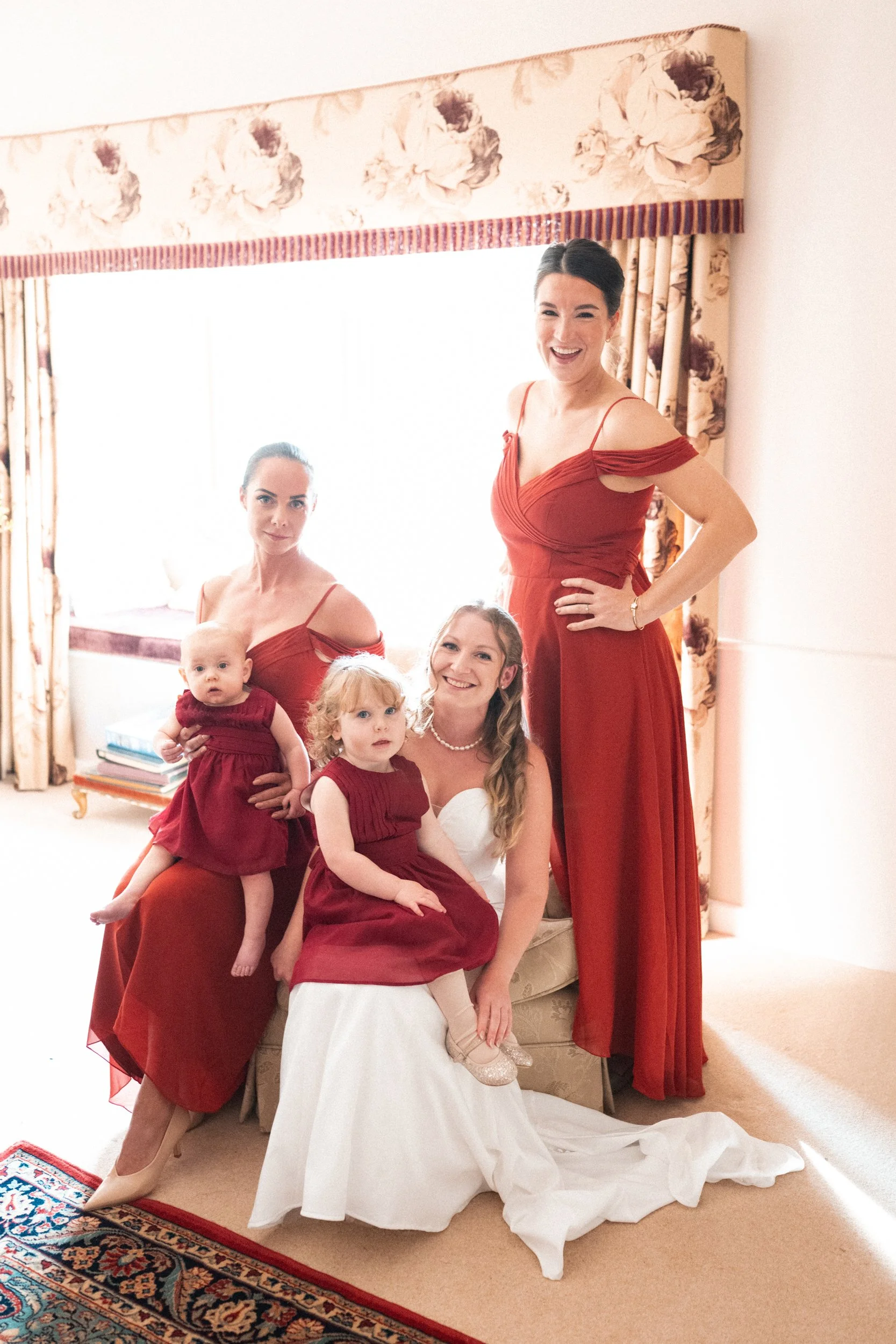 Group of women and two children in a bright room with floral curtains, celebrating a wedding; the woman in the center wears a white wedding dress, lesurely sitting on a sofa; others in red dresses around her.