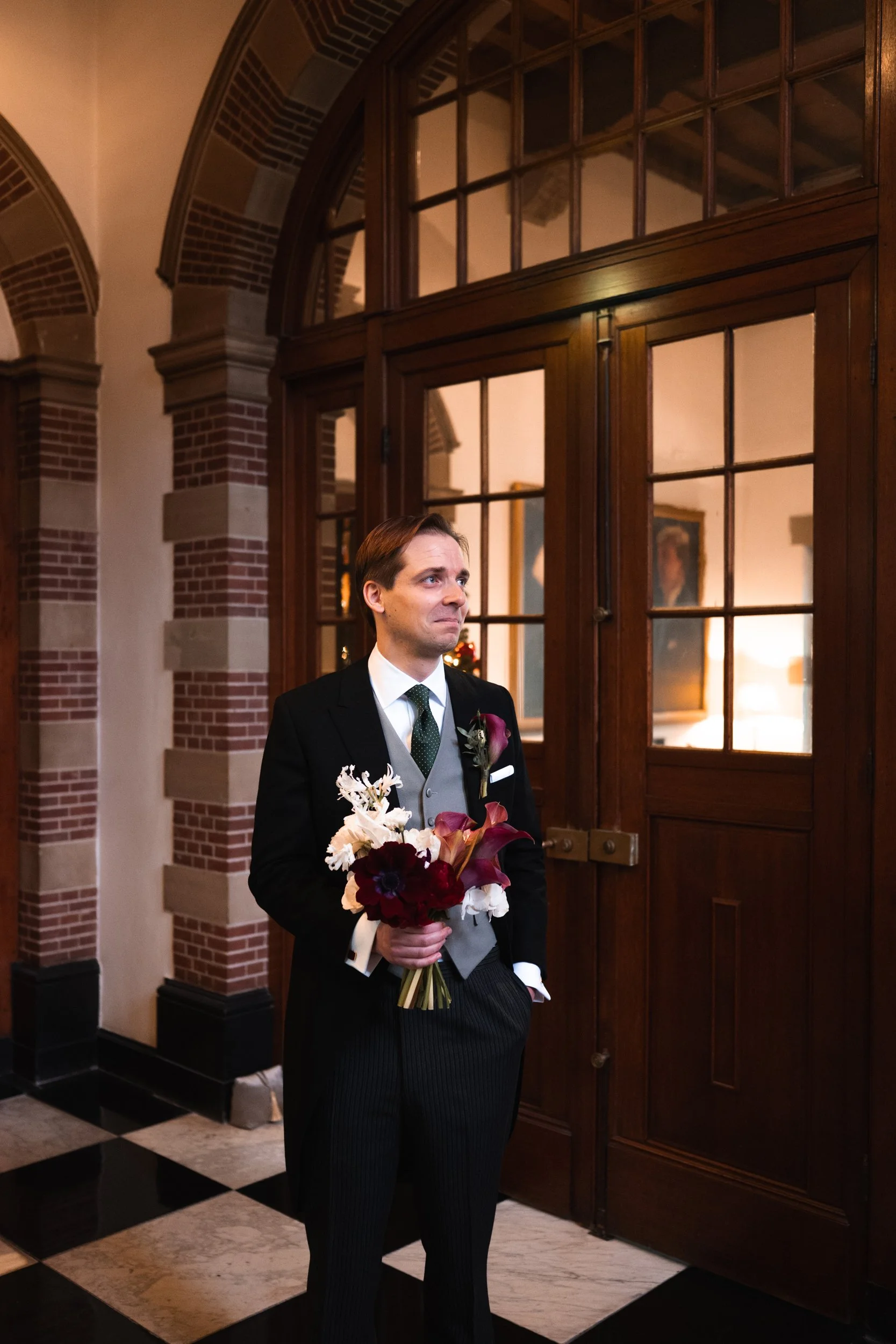 A man in formal attire holding a bouquet of flowers, standing indoors near wooden double doors with glass panels, in a setting with brick and stone architecture.