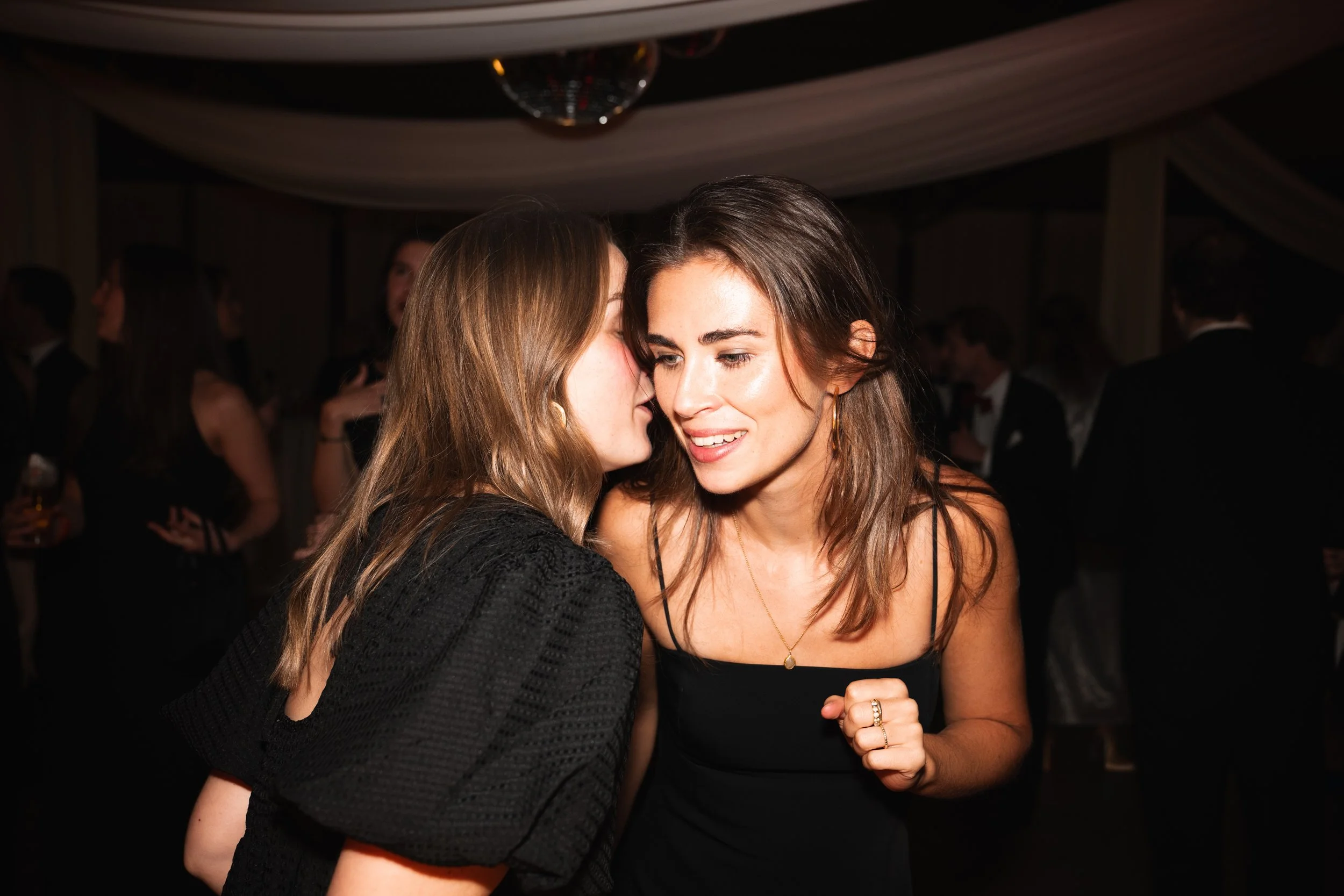 Two women talk softly at a social event, surrounded by other guests in formal attire.