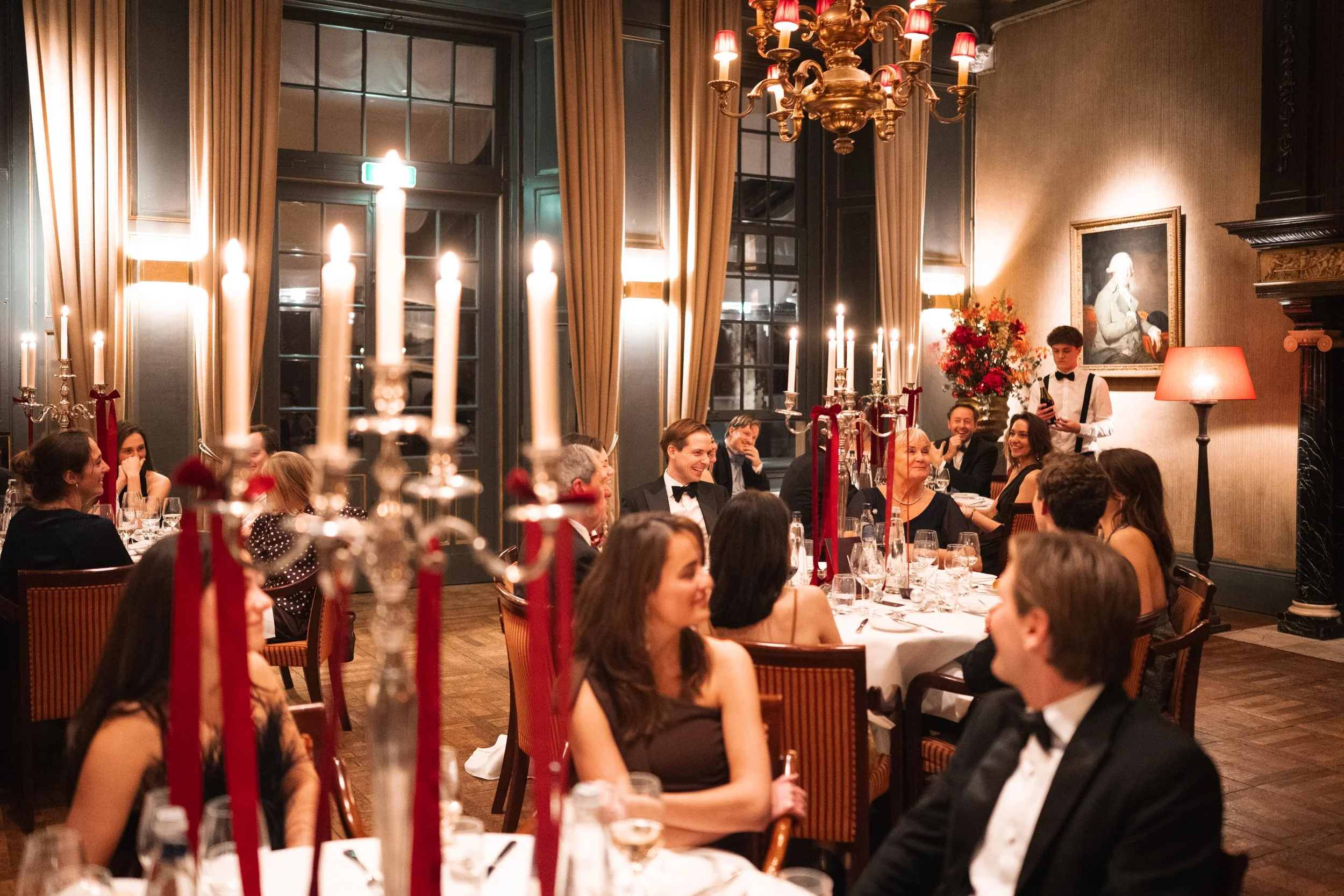 Guests at a formal dinner party in an elegant dining room with chandeliers, candelabras, and framed artwork, enjoying a festive celebration.