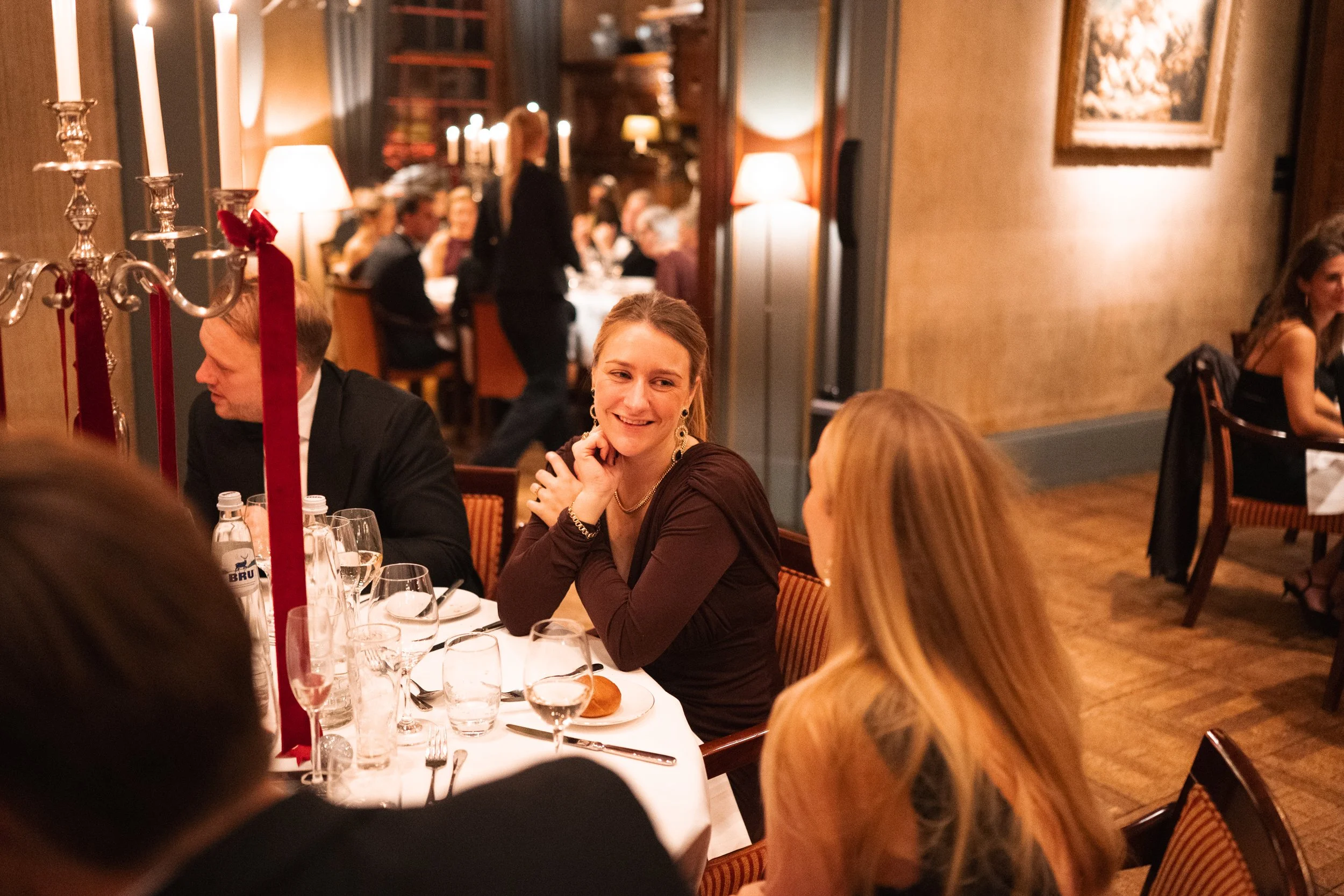 People dining at a restaurant, including a woman with earrings and a woman with long blonde hair talking to her, others in the background, warm lighting, and a candelabra with red ribbons.