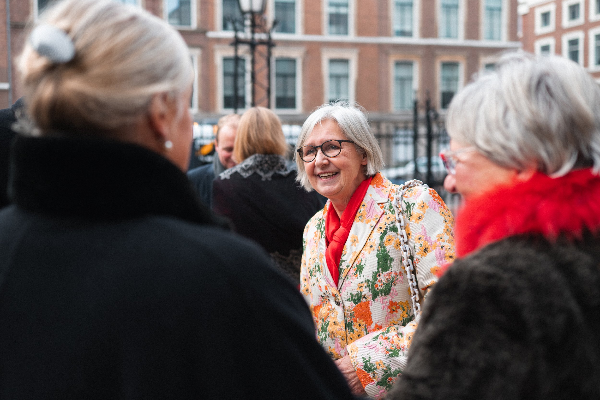 Four older women talking outdoors in an urban setting, with brick buildings in the background. One woman wears a floral blazer and red scarf, smiling at another woman in black with a gray hair clip.