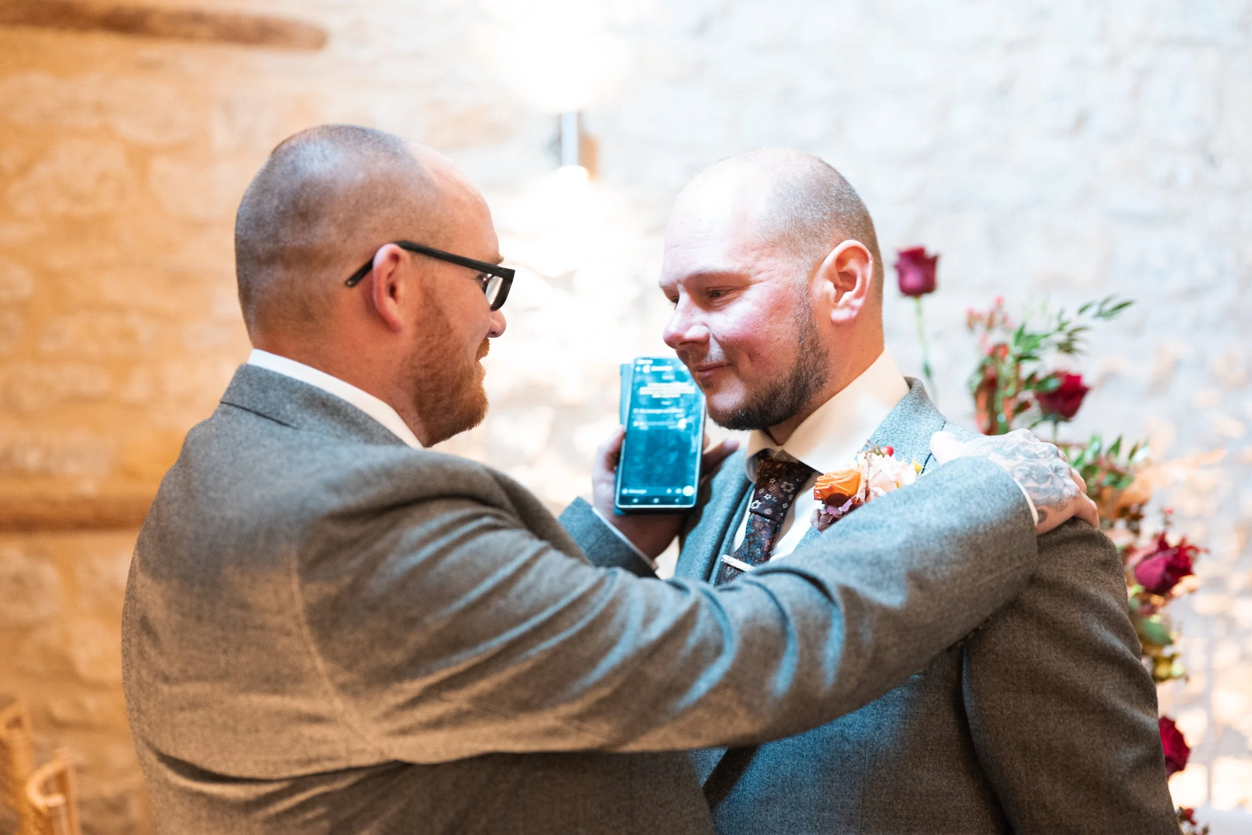 Two men in suits at a wedding, one is passing a phone to the other during a ceremony with a brick wall and flower arrangement in the background.