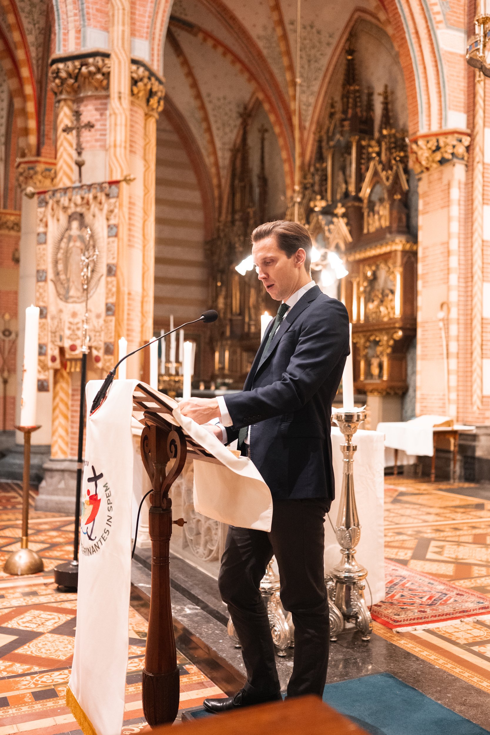 A man in a suit reading from a lectern inside a church with ornate gold decorations, candles, and religious symbols.