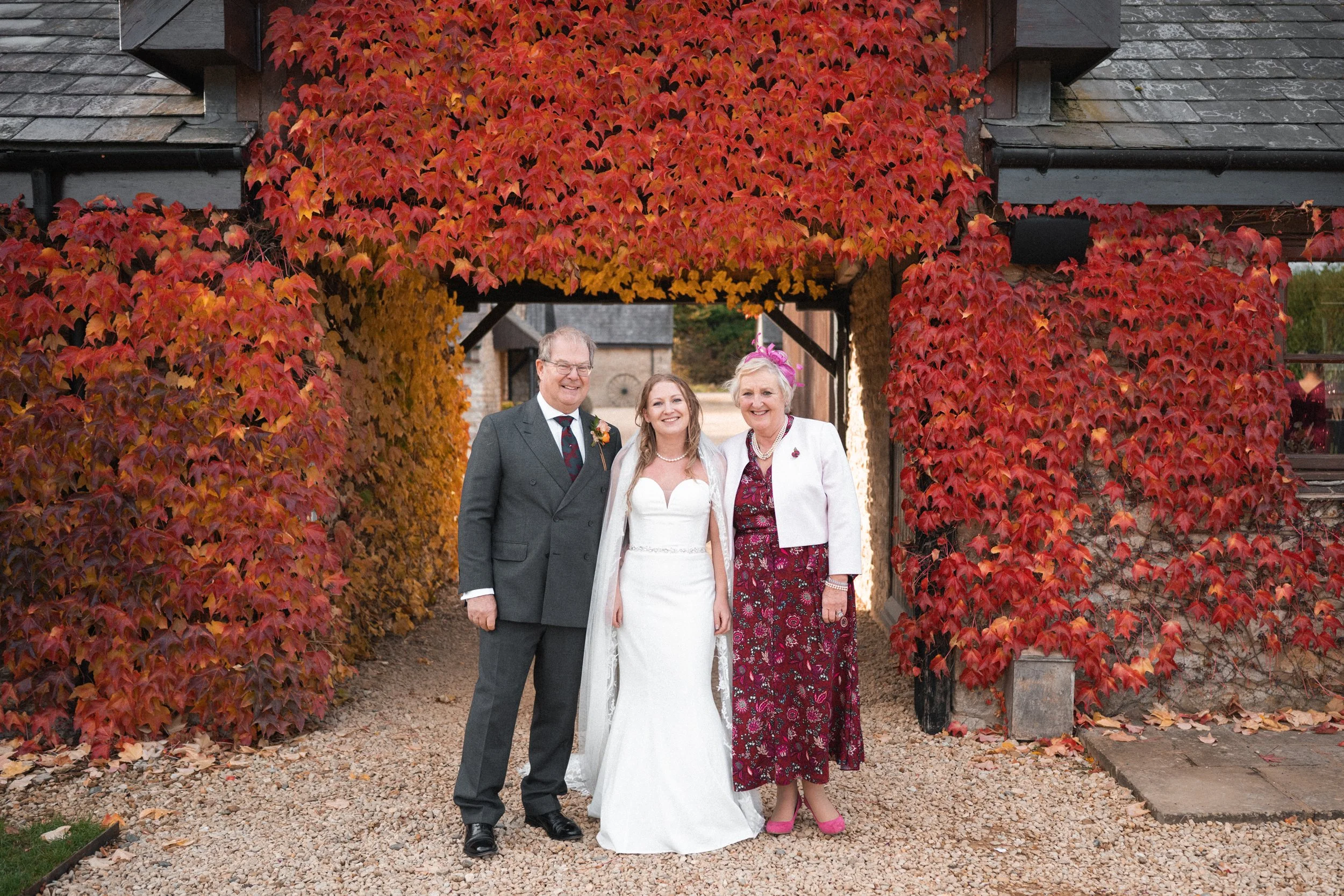 A bride in a white wedding dress stands between a man in a gray suit and a woman in a floral dress and white jacket, under an archway covered in red and orange autumn leaves, on a gravel path.