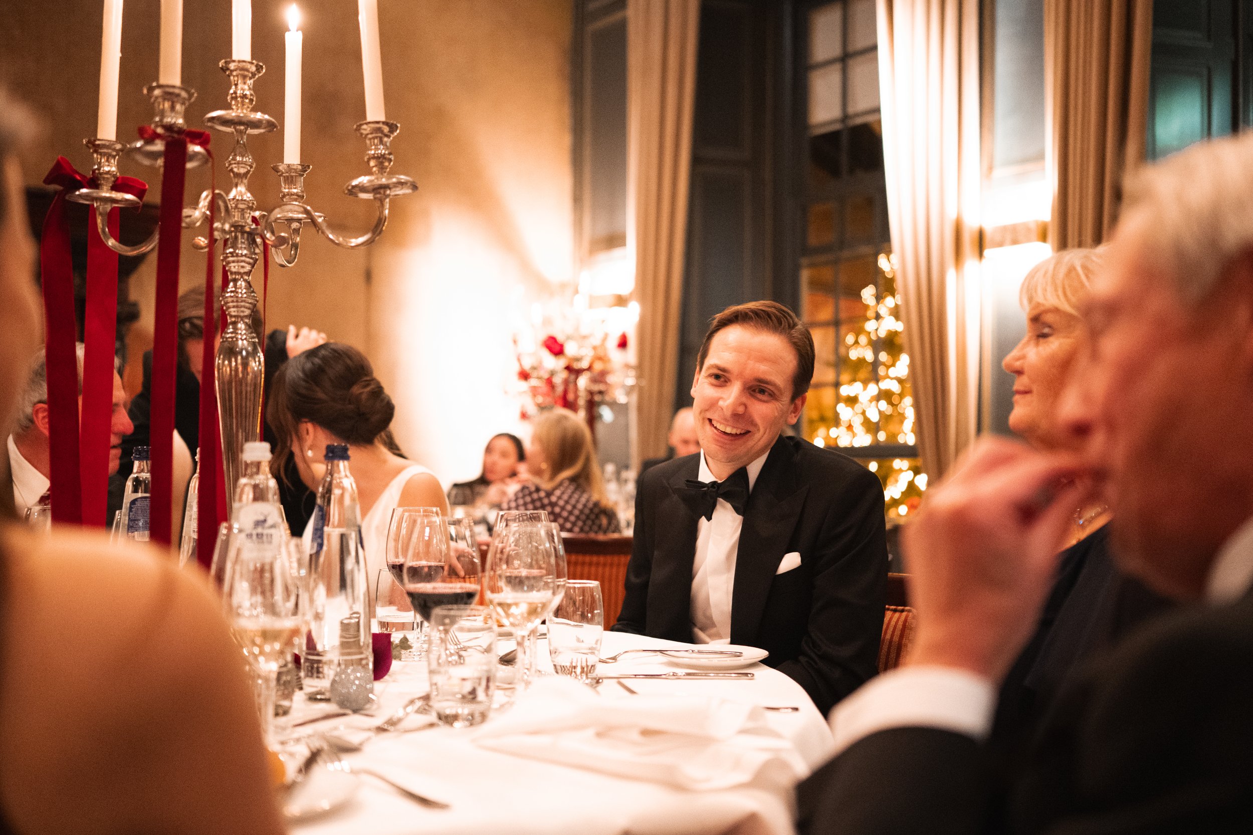 A man in a tuxedo smiling and talking to an older woman at a formal dinner table decorated with candles, wine glasses, and bottles, in an elegant room with warm lighting and large windows.