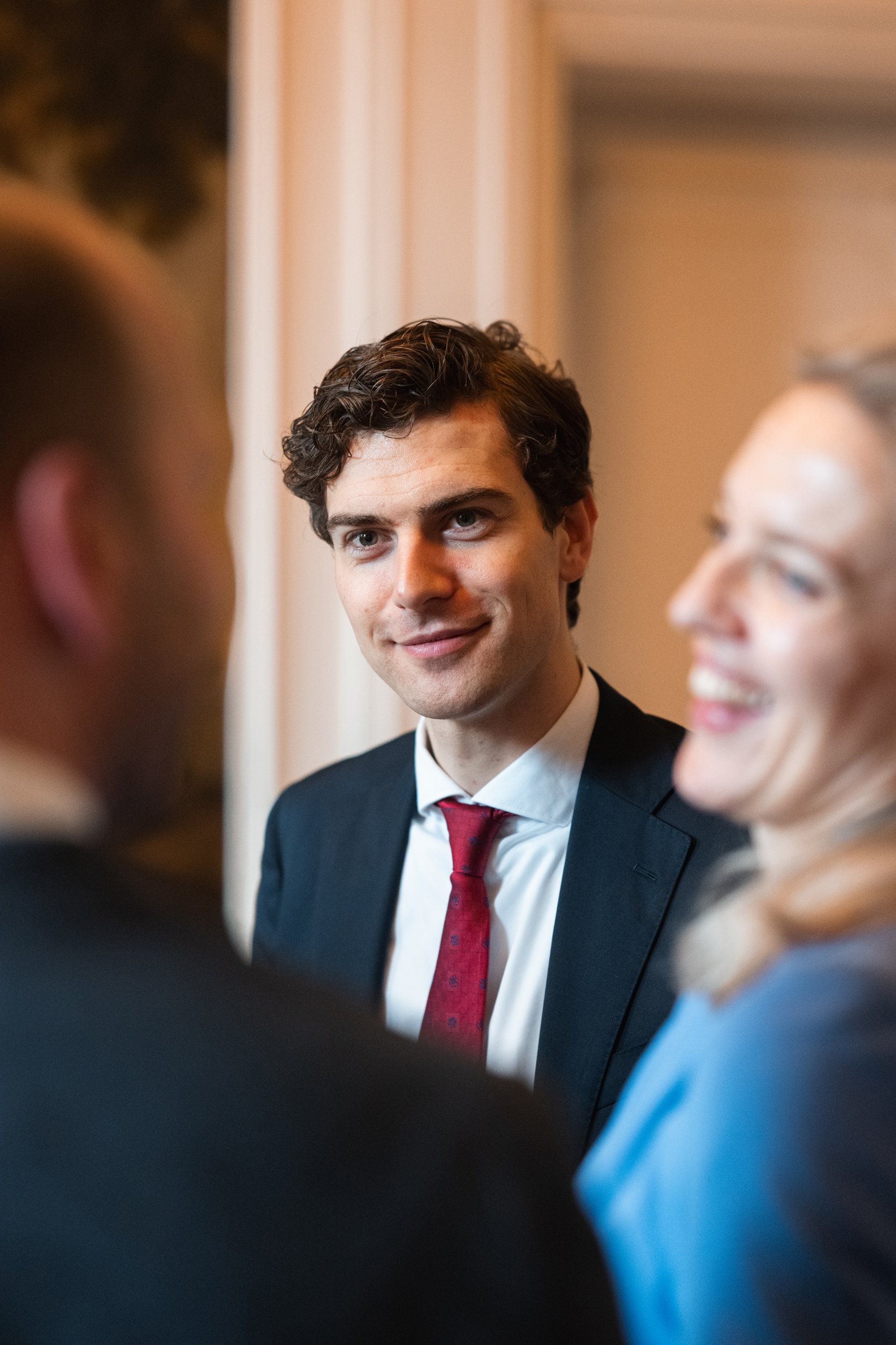 A young man in a suit with a red tie smiling and looking at someone, with two blurred people in the foreground.