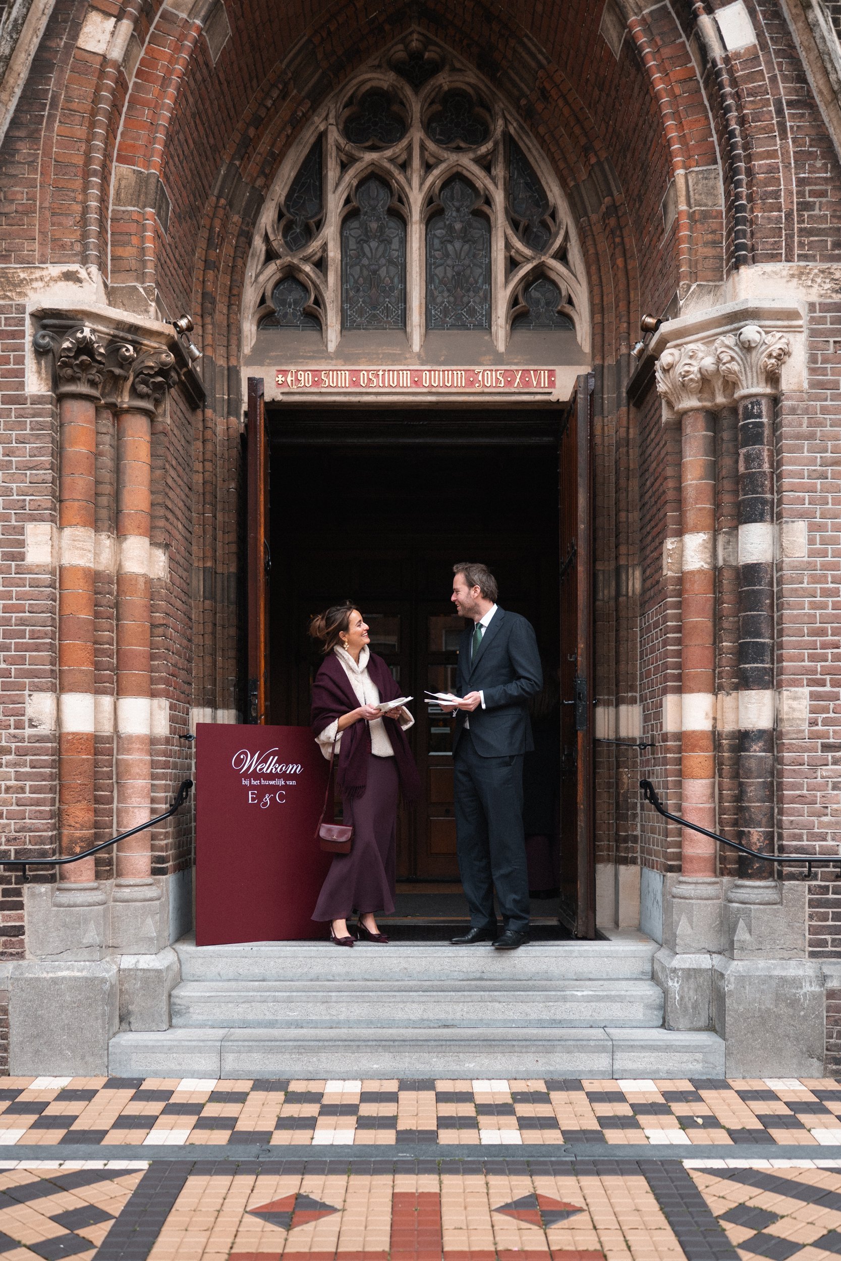 A man and woman standing on the steps of a church engaged in conversation, with the man holding papers, both dressed in formal attire, and the woman dressed in a purple coat and scarf. The church features brick architecture with decorative stone acce
