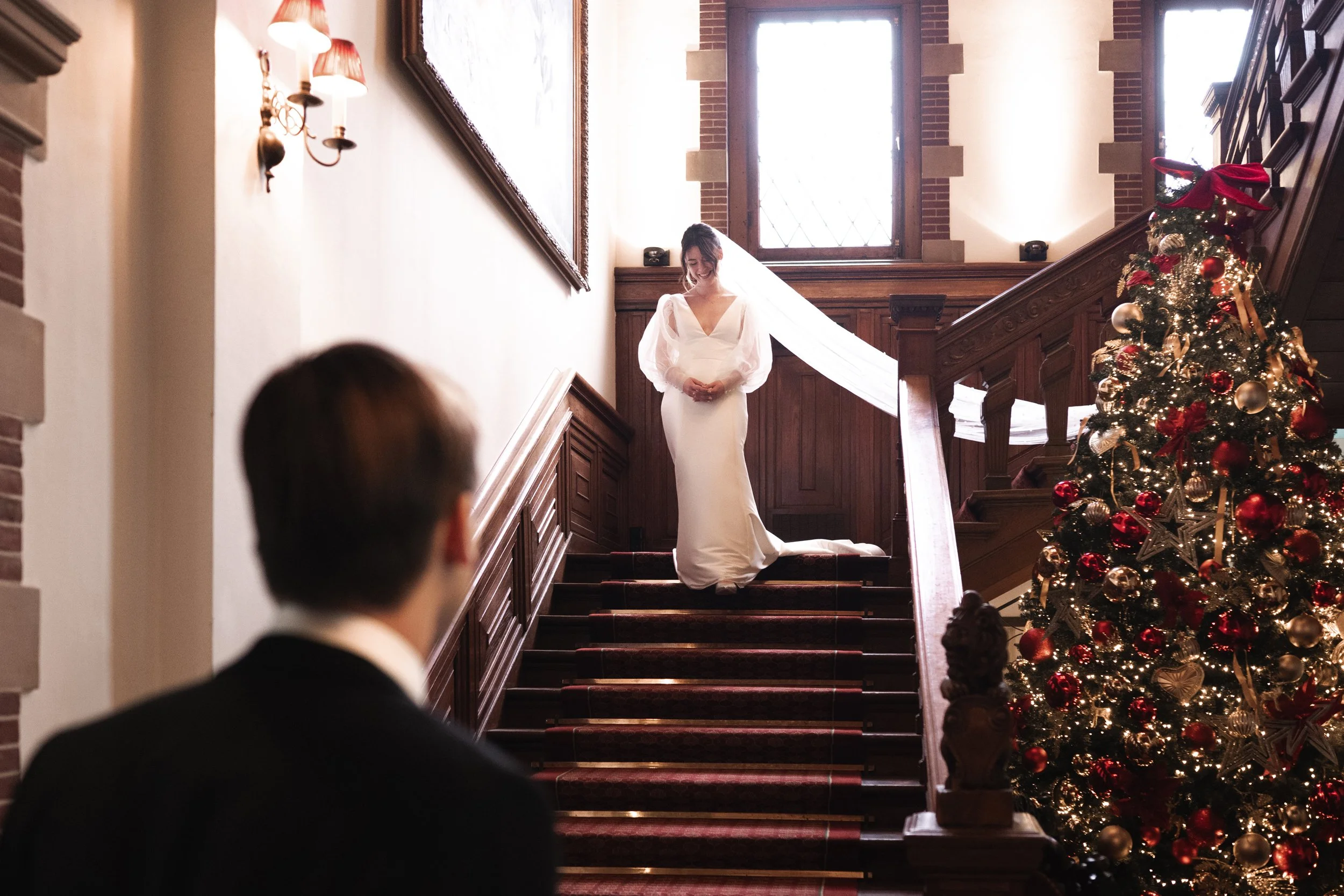 A bride in a white wedding dress standing on a stairway, looking down at a groom in a black suit, with a decorated Christmas tree nearby in a warmly lit interior space.