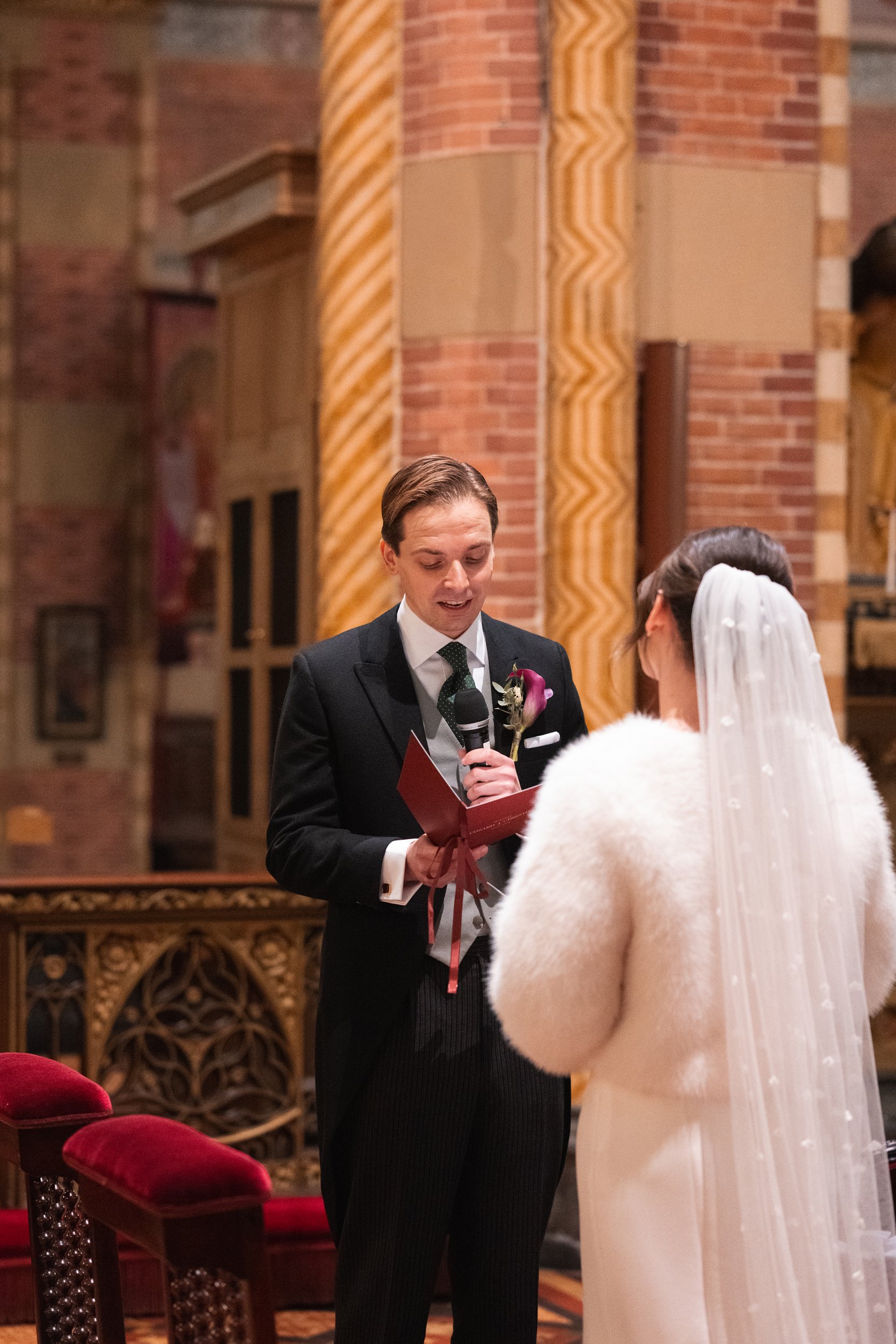 A groom and bride exchange vows during a wedding ceremony inside a church, with the groom holding a red book and speaking into a microphone.