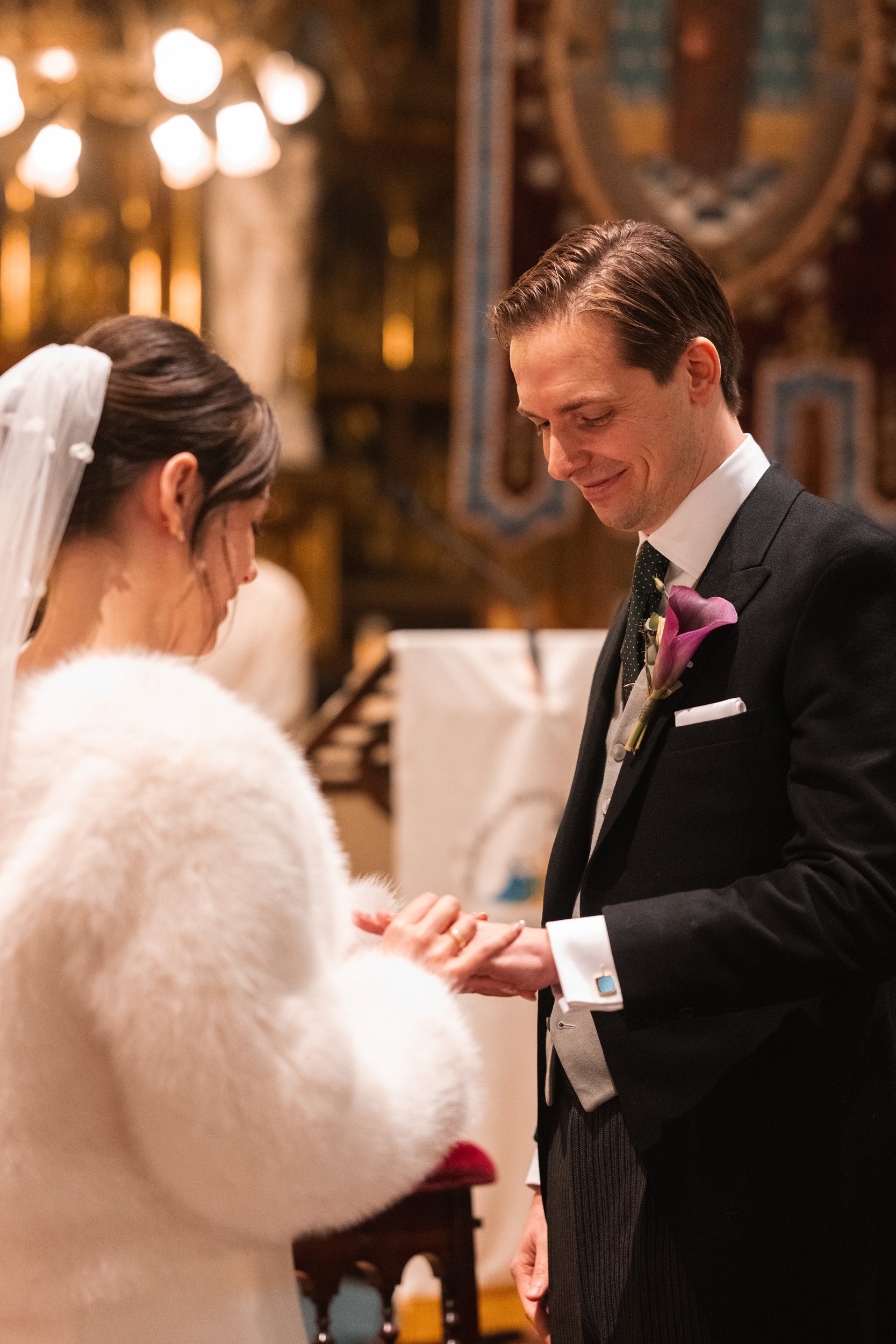 A bride and groom during their wedding ceremony, holding hands and smiling in a church with warm lighting and ornate decorations.
