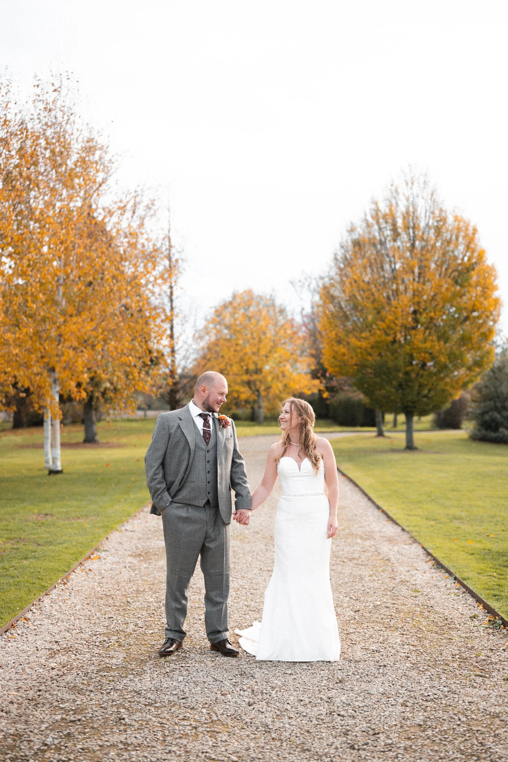 A newlywed couple walking hand in hand on a gravel path in a park with autumn trees with orange leaves.
