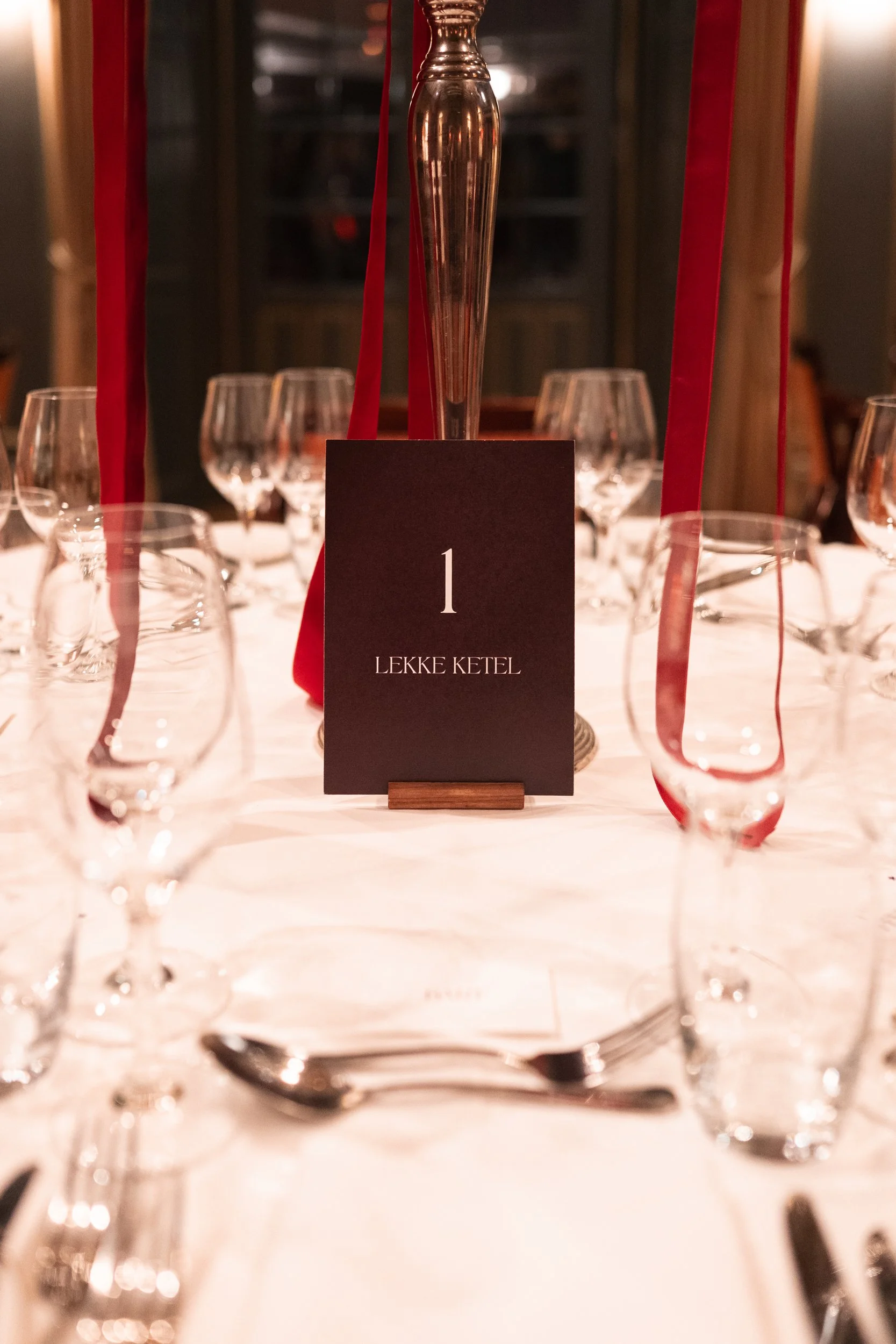 Elegant dining table setup with a black table number card reading '1 Leke Ketel' in the center, surrounded by multiple wine glasses, water glasses, and silverware, with red ribbons hanging from a tall, decorative centerpiece.