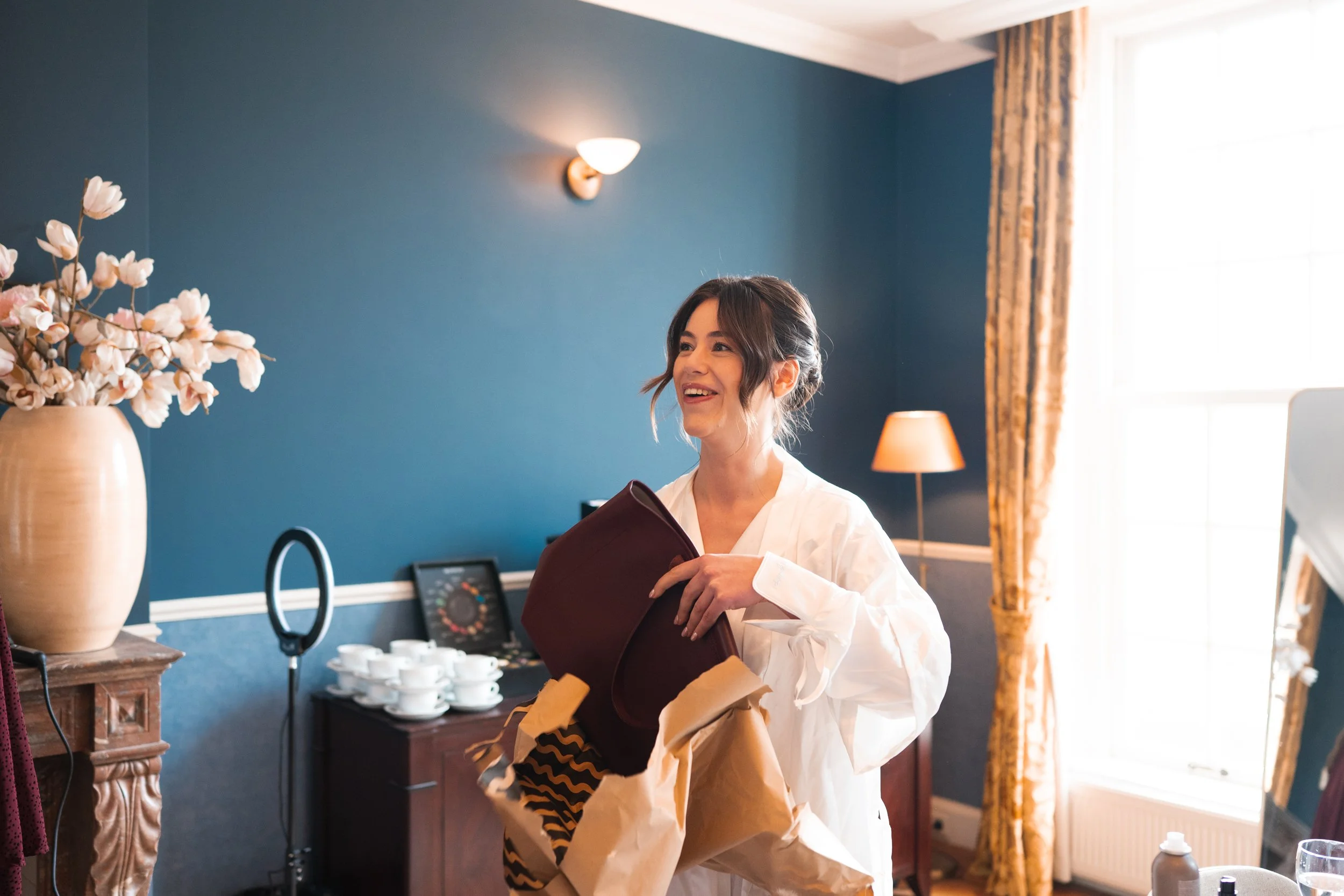 A woman with dark hair in a white robe is smiling while holding a brown handbag and gift bag in a room with blue walls, a large window with yellow curtains, a side table with stacked white cups, a vase with flowers, and a lamp.