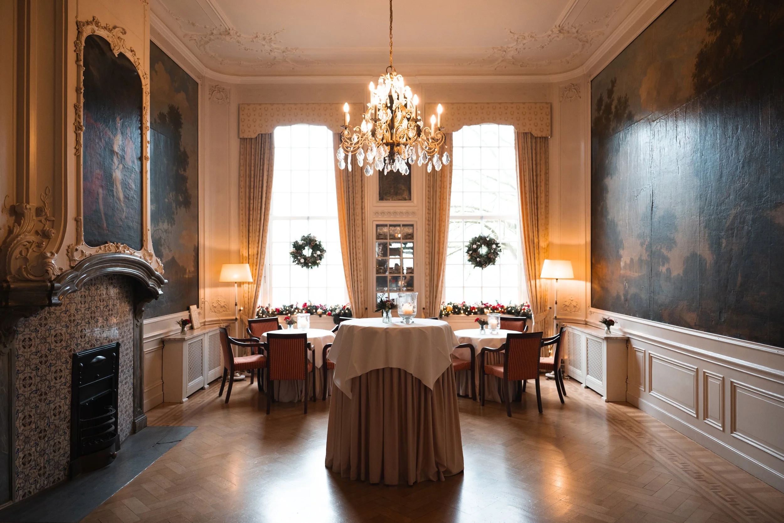 Elegant, decorated dining room with a chandelier, large windows with curtains, round tables with white tablecloths, chairs, holiday wreaths, and floral arrangements, with festive decorations and warm lighting.