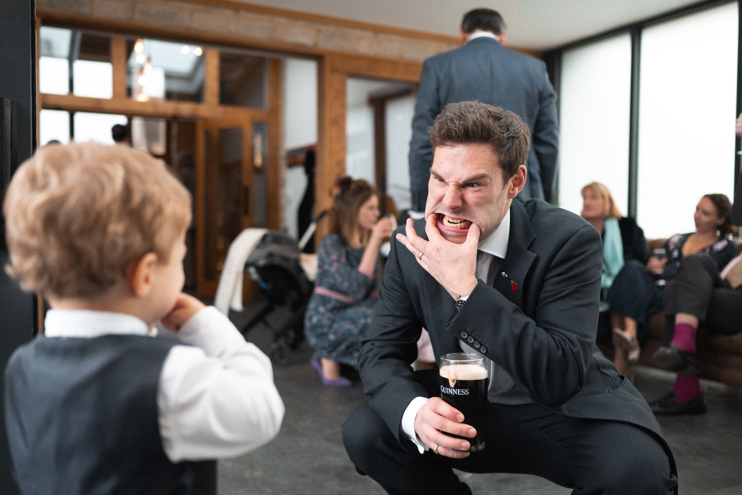 A man in a suit making a grimace and biting his finger while holding a pint of Guinness, sitting face to face with a young boy in a formal vest at a social gathering.