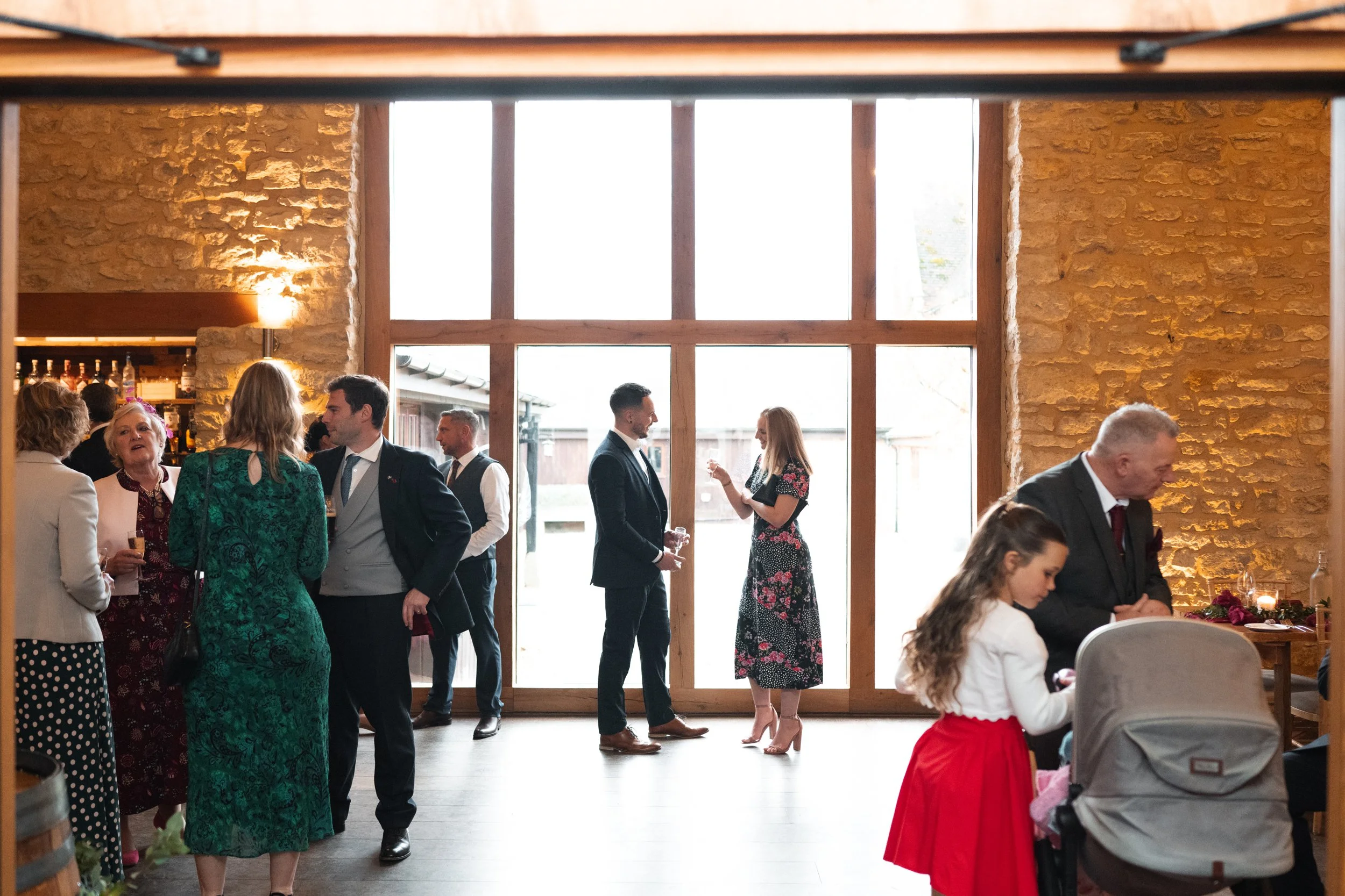 People socializing at an indoor event with large windows, brick walls, and a bar in the background.