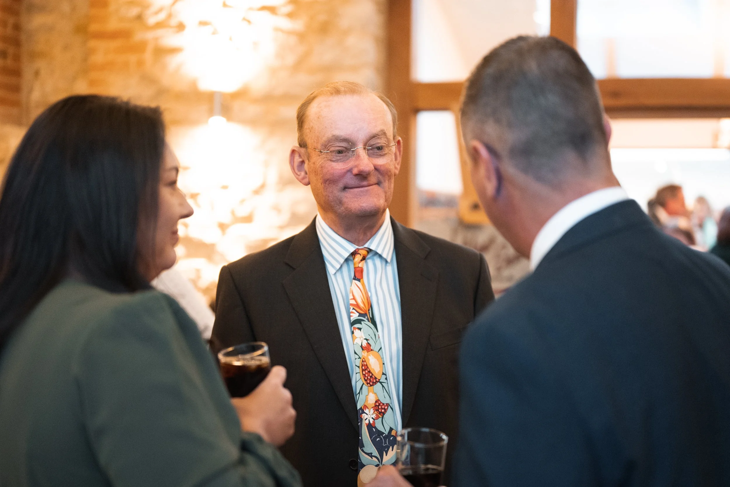 Three people engaged in conversation at a social event, with a brightly lit brick wall and window in the background.