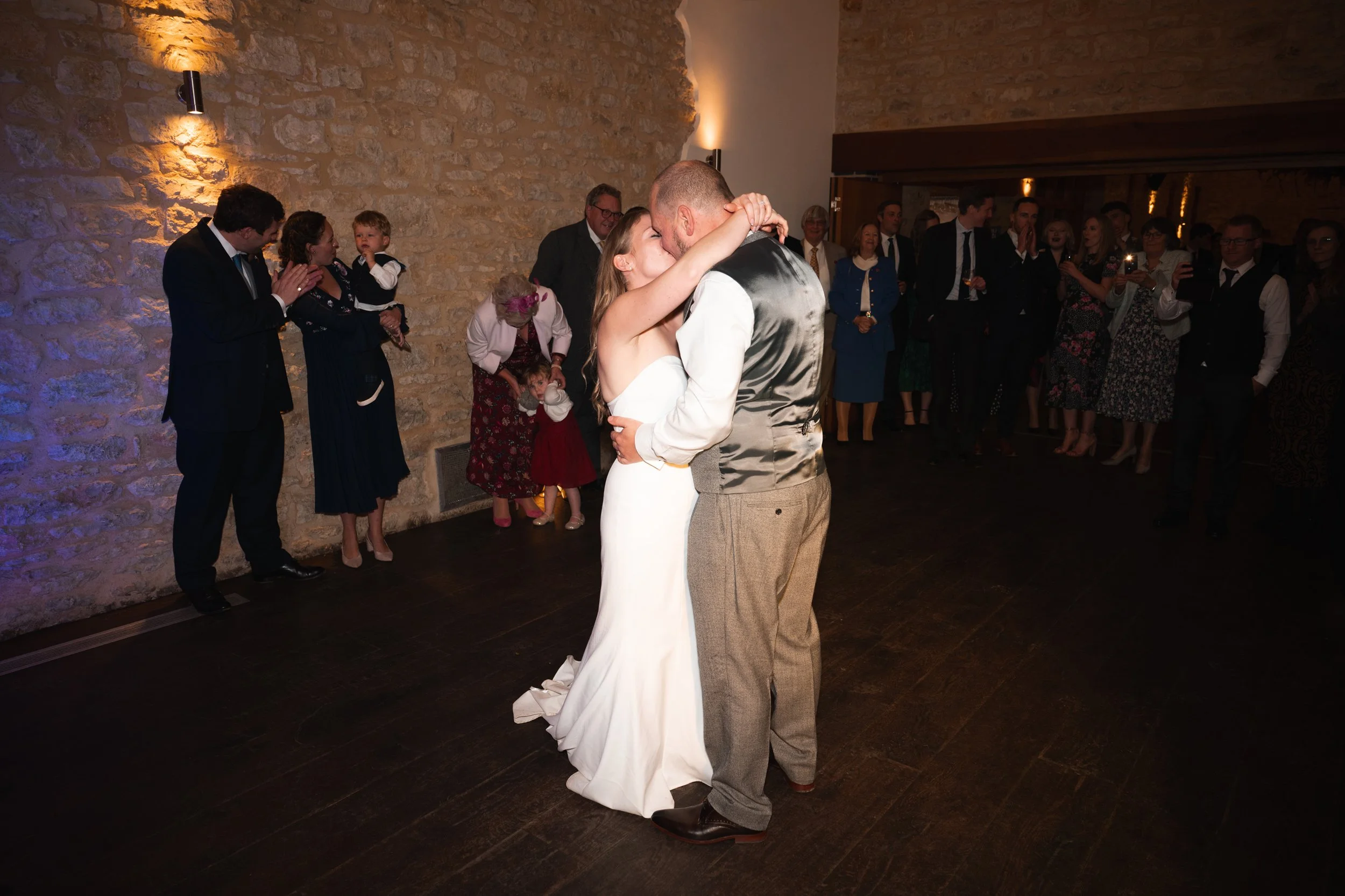 A bride and groom sharing a dance at their wedding reception, surrounded by guests in a warmly lit room with stone walls.