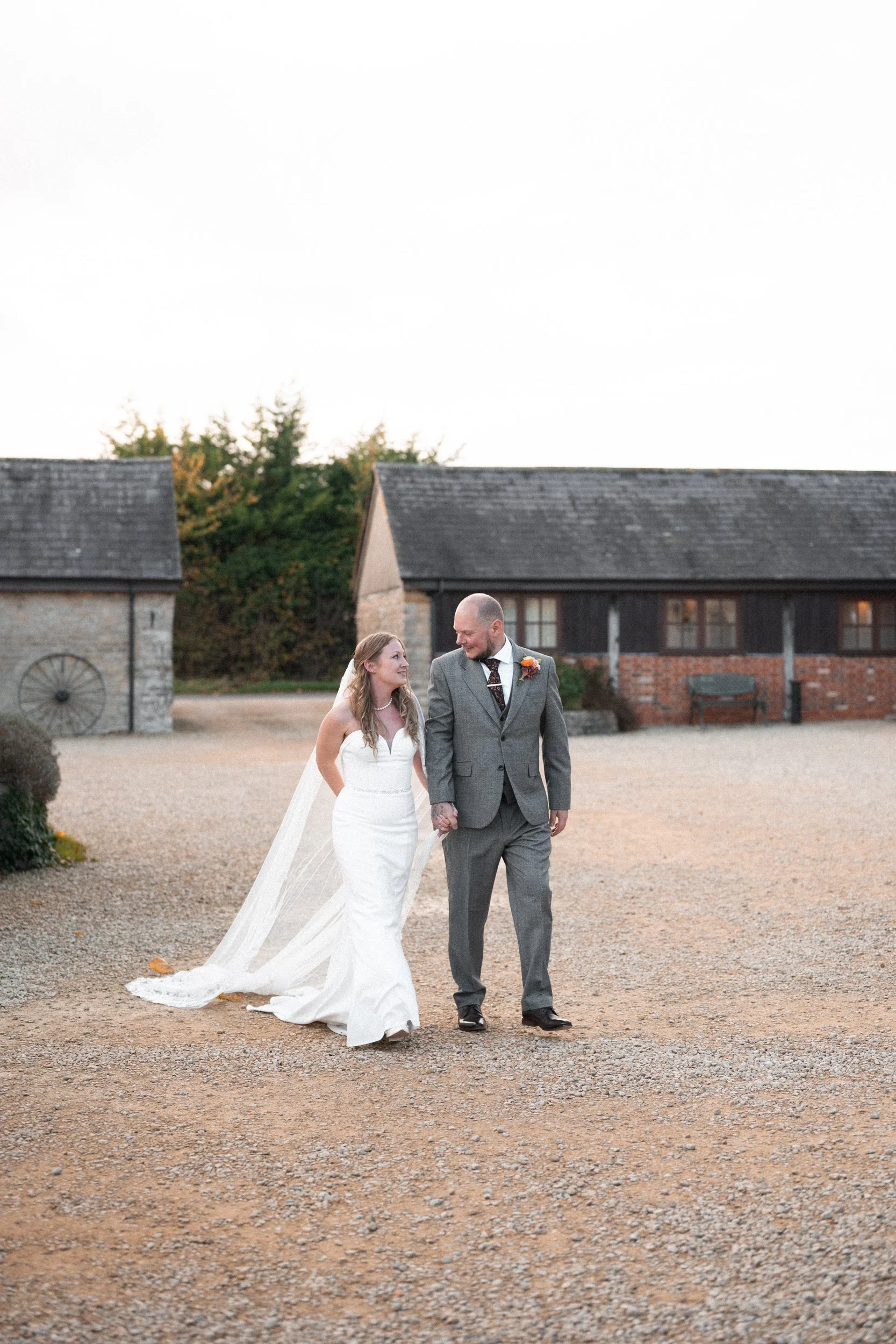 A bride and groom walking hand in hand on a gravel path, smiling at each other during their wedding day outside with rustic buildings in the background.