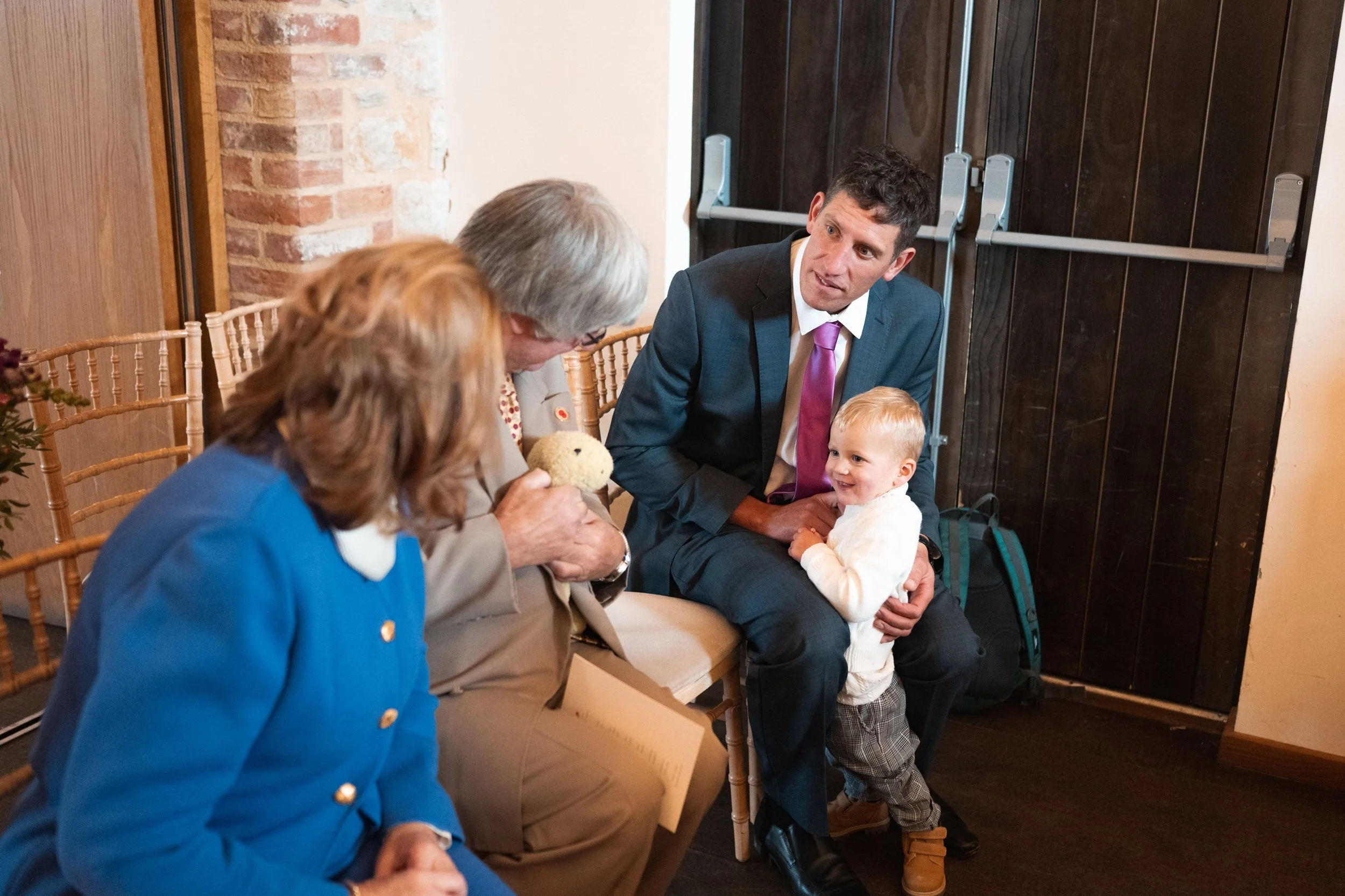 A man in a suit sits holding a young boy, while an elderly woman holds a teddy bear and interacts with two women dressed in blue and tan, in a cozy room with brick and wooden walls.