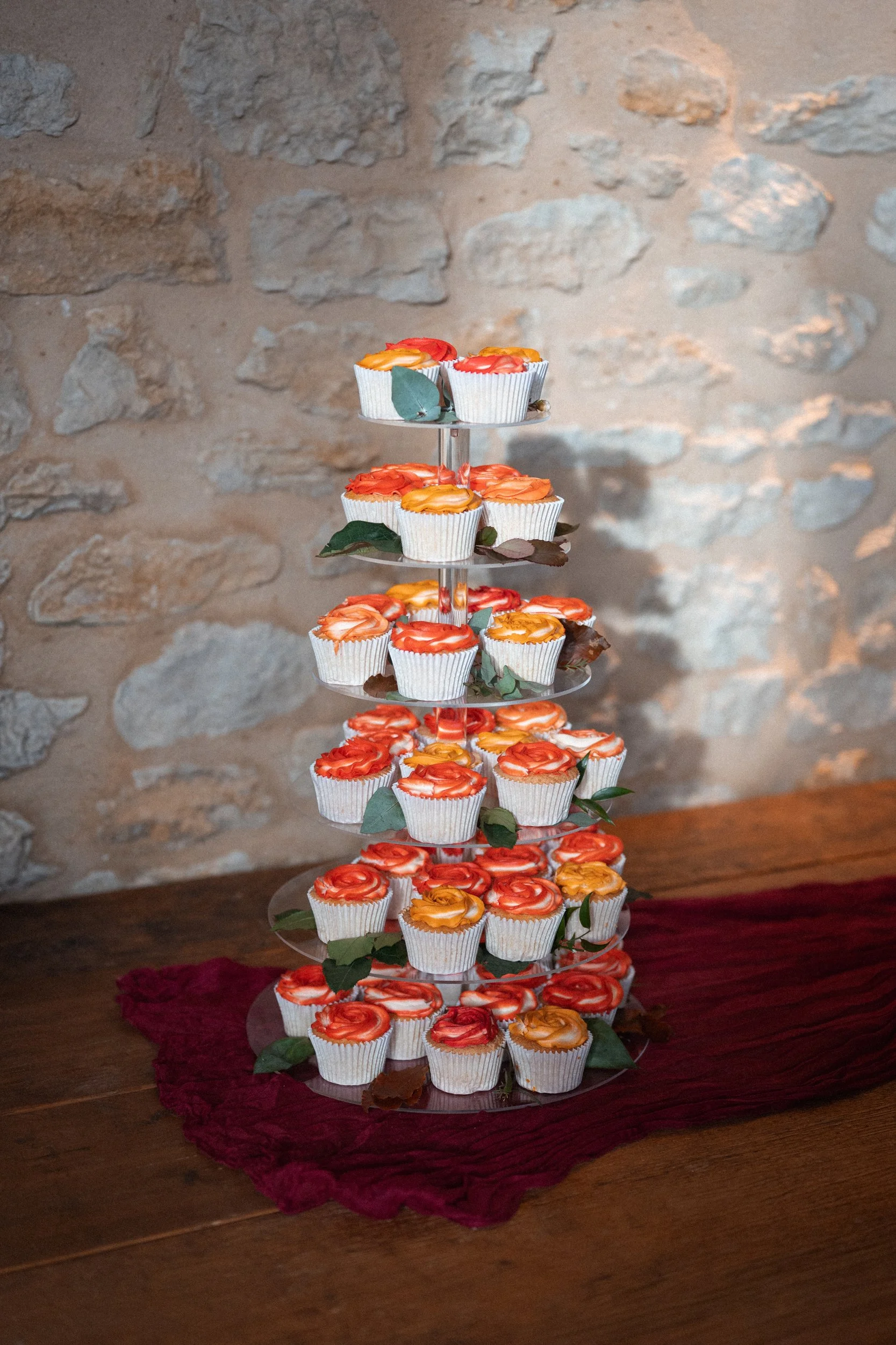 A tiered stand with decorated cupcakes on a wooden table, against a stone wall background.