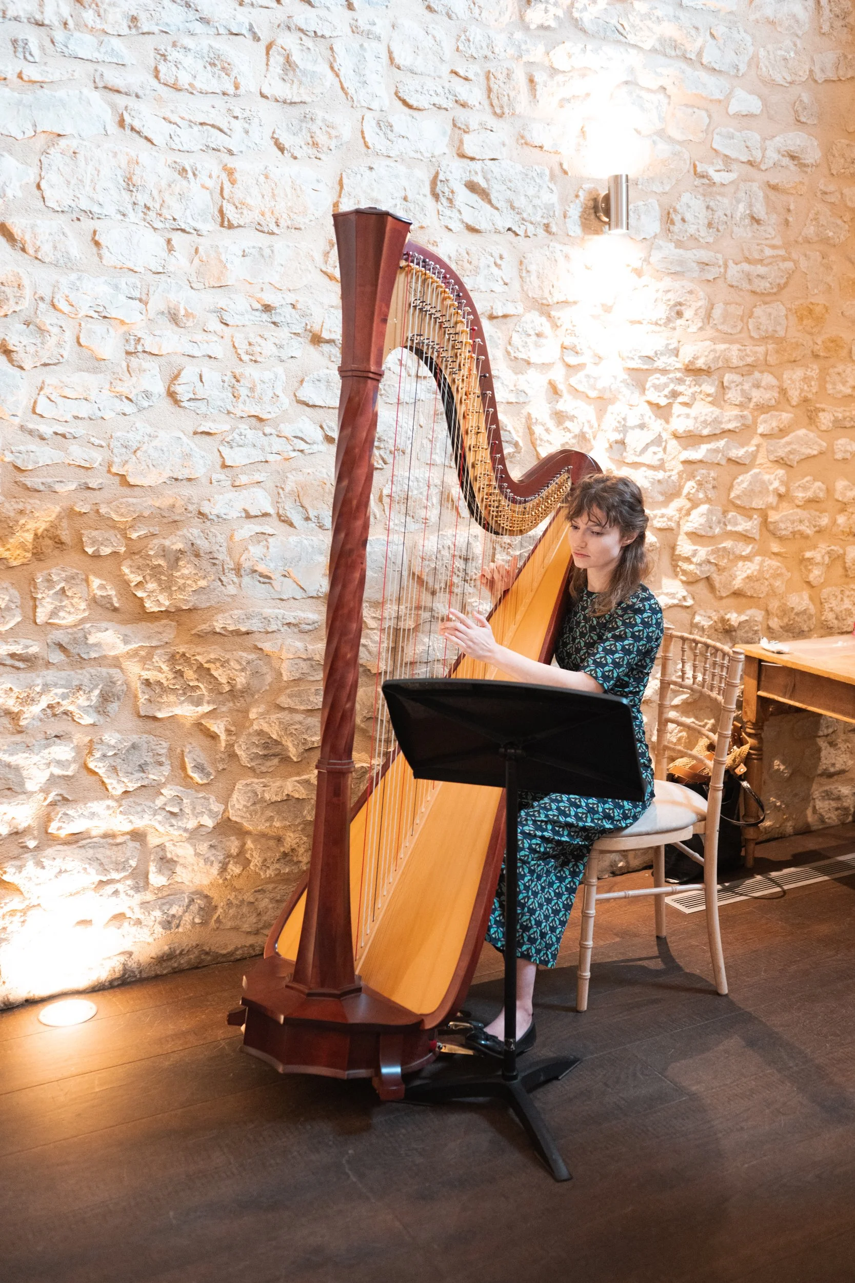 A woman sitting on a white chair playing a large wooden harp in a room with a stone wall background.
