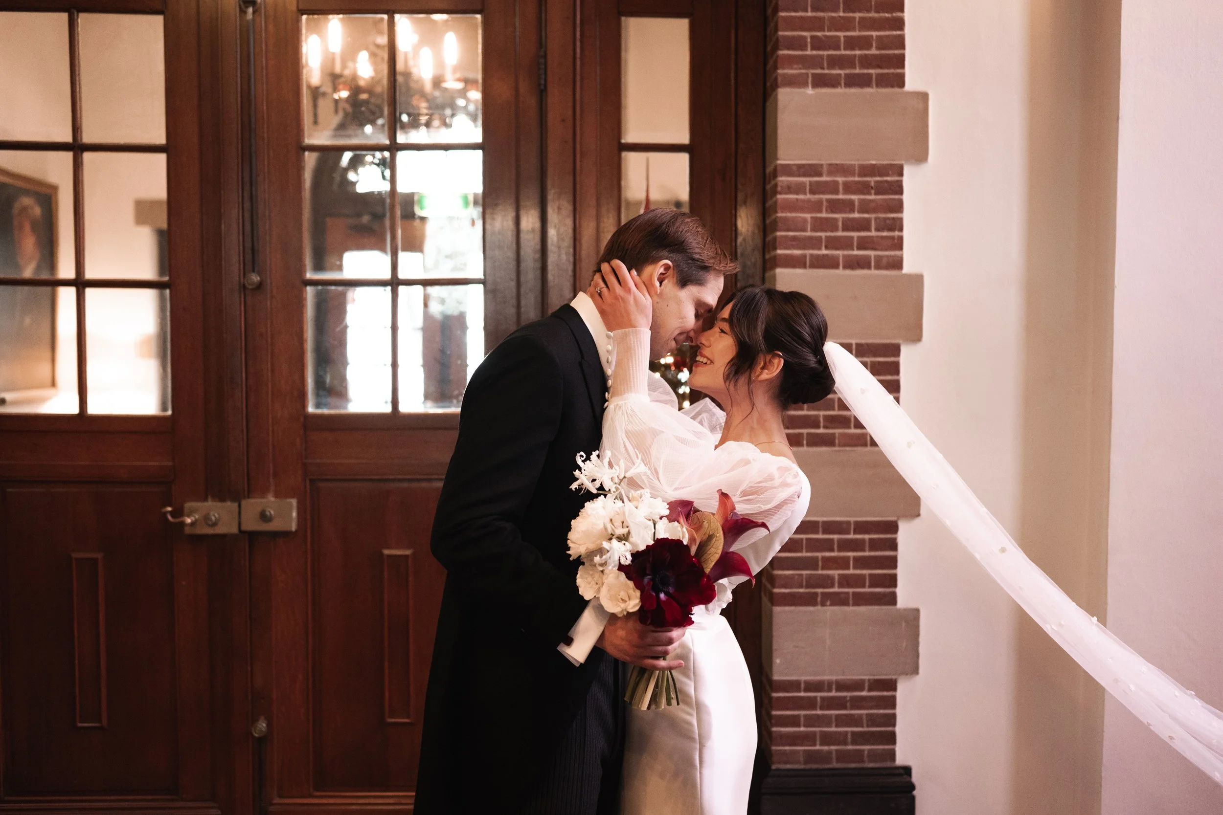 A bride and groom in wedding attire sharing a close, joyful moment indoors, with the groom holding a bouquet of red and white flowers.