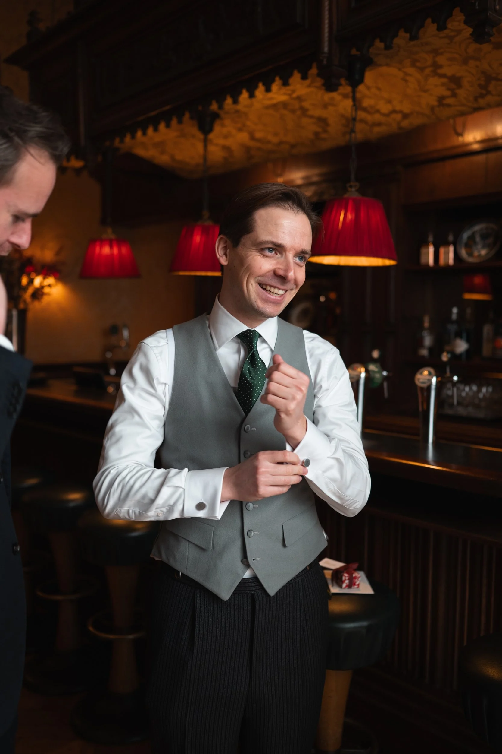 A man in a gray vest, white shirt, and dark tie smiling and gesturing while talking in a dimly lit bar with red lamps and dark wooden decor.