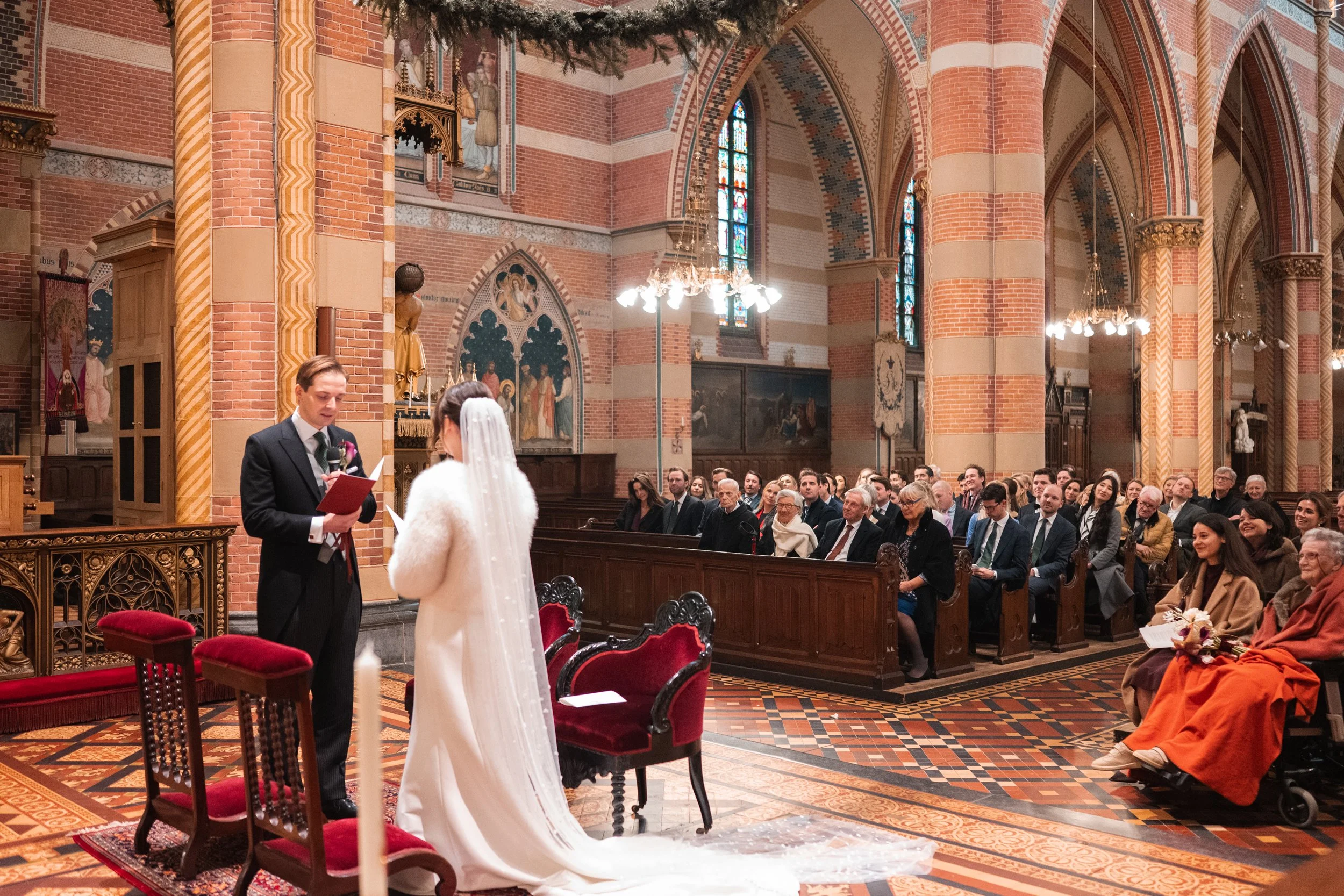 A wedding ceremony inside a church with high vaulted ceilings, stained glass windows, and ornate artwork. A groom in a black suit is reading vows to a bride in a white wedding gown with a veil. Guests are seated in pews, watching the couple exchange 
