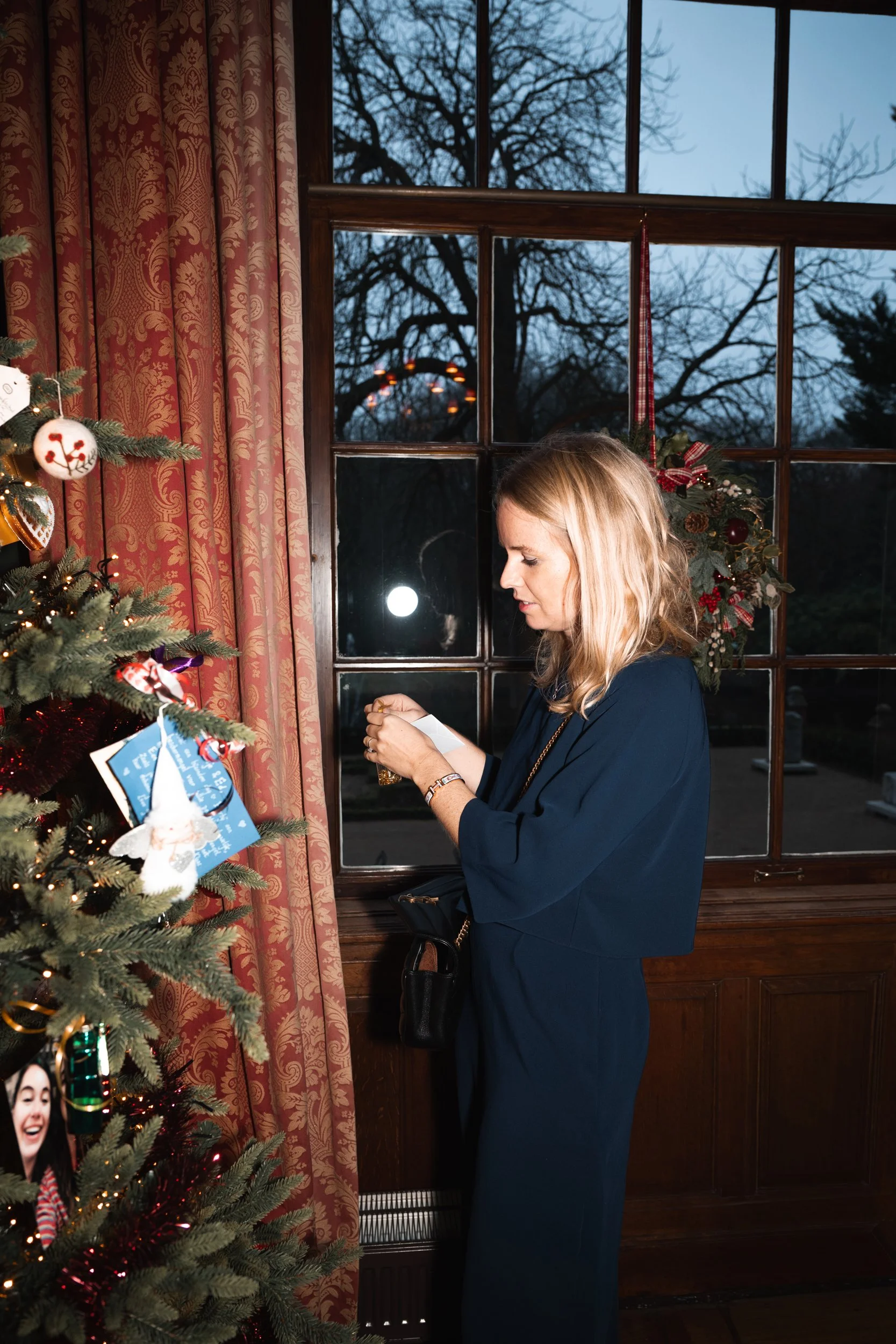 A woman in a blue dress standing beside a decorated Christmas tree, holding an ornament and a piece of paper, in a room with red patterned curtains and a window showing trees and an overcast sky.