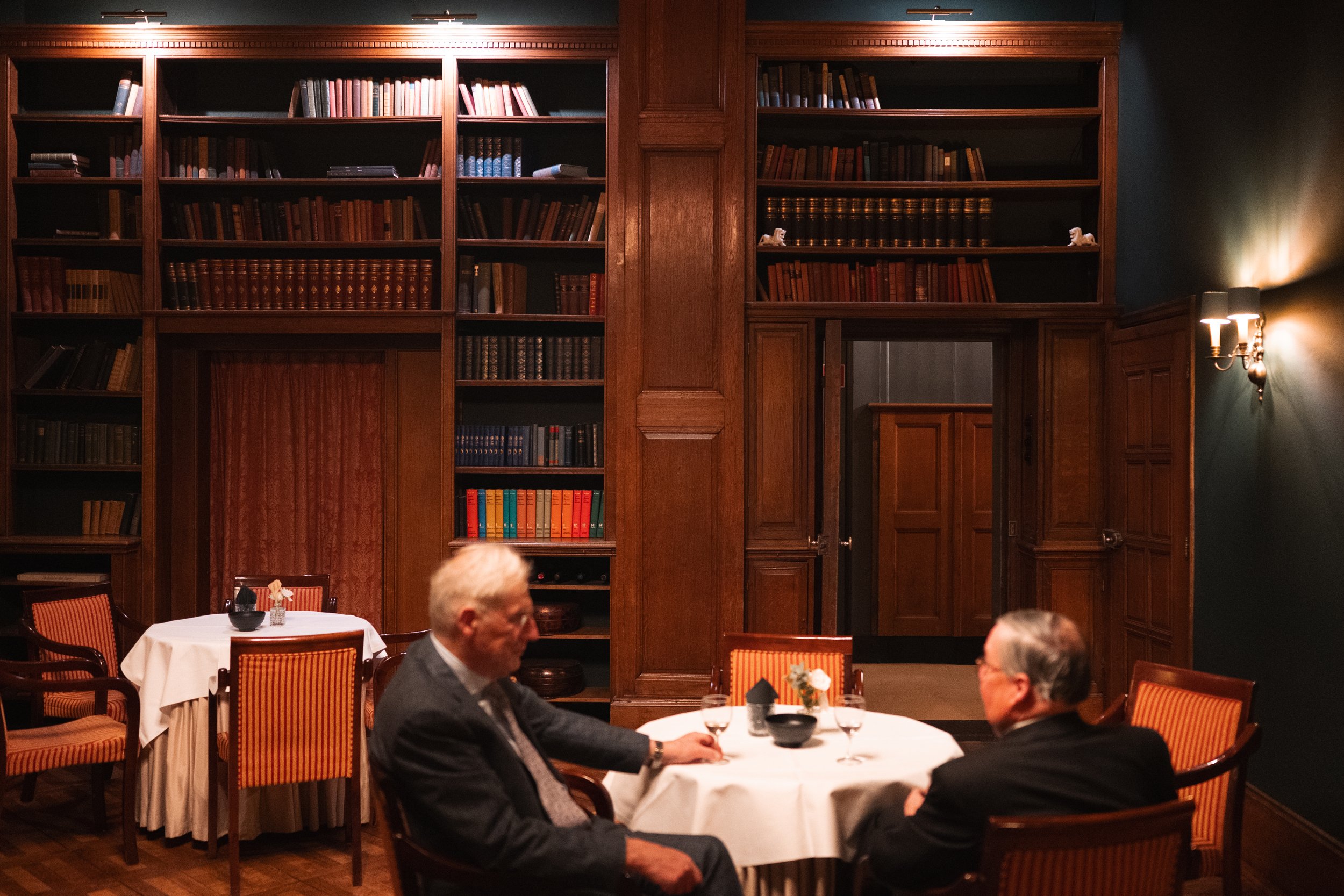 Two older men in suits sitting at a round table with white cloth in a wood-paneled library or private dining room, with bookshelves filled with books in the background, dim lighting, and flower arrangements on the tables.