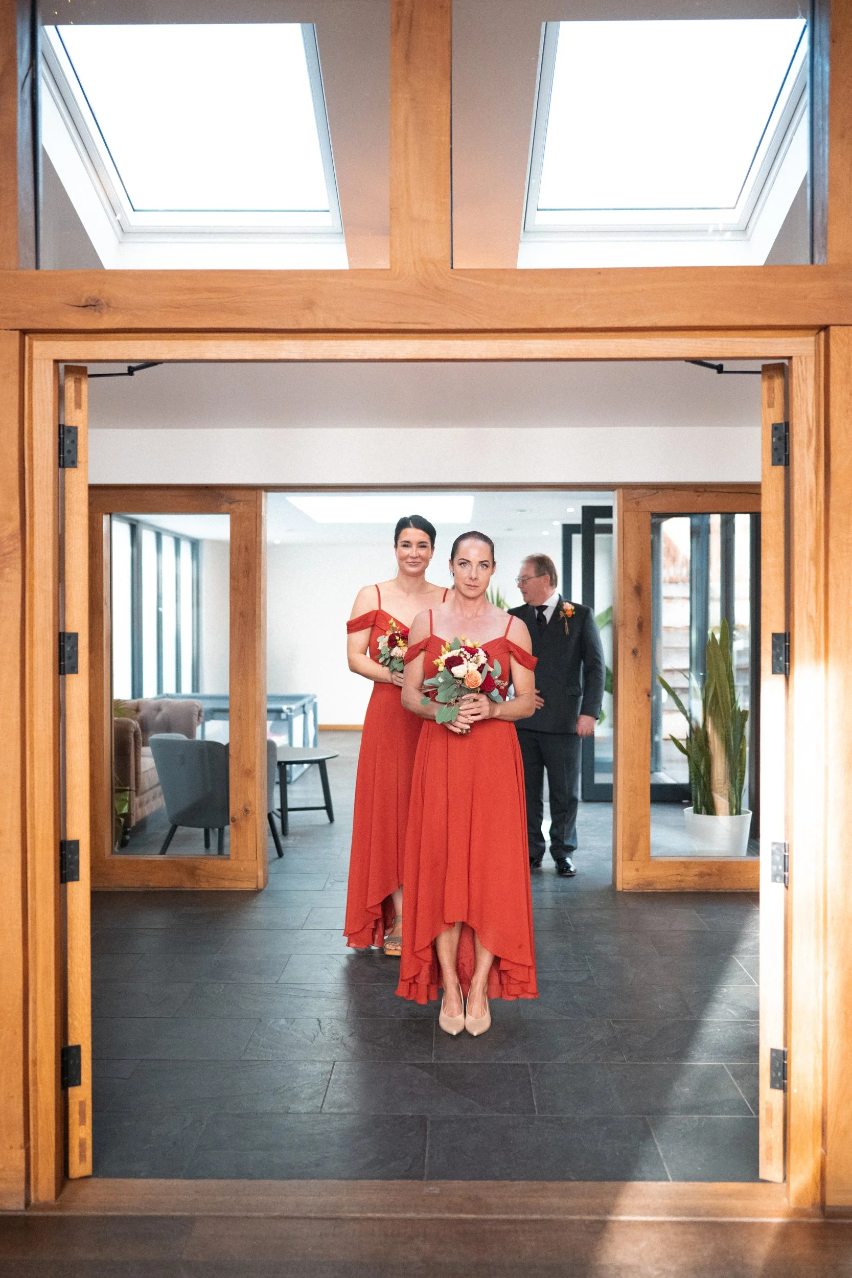 Three women in red dresses holding bouquets in a modern indoor setting, with a man in a tuxedo in the background.