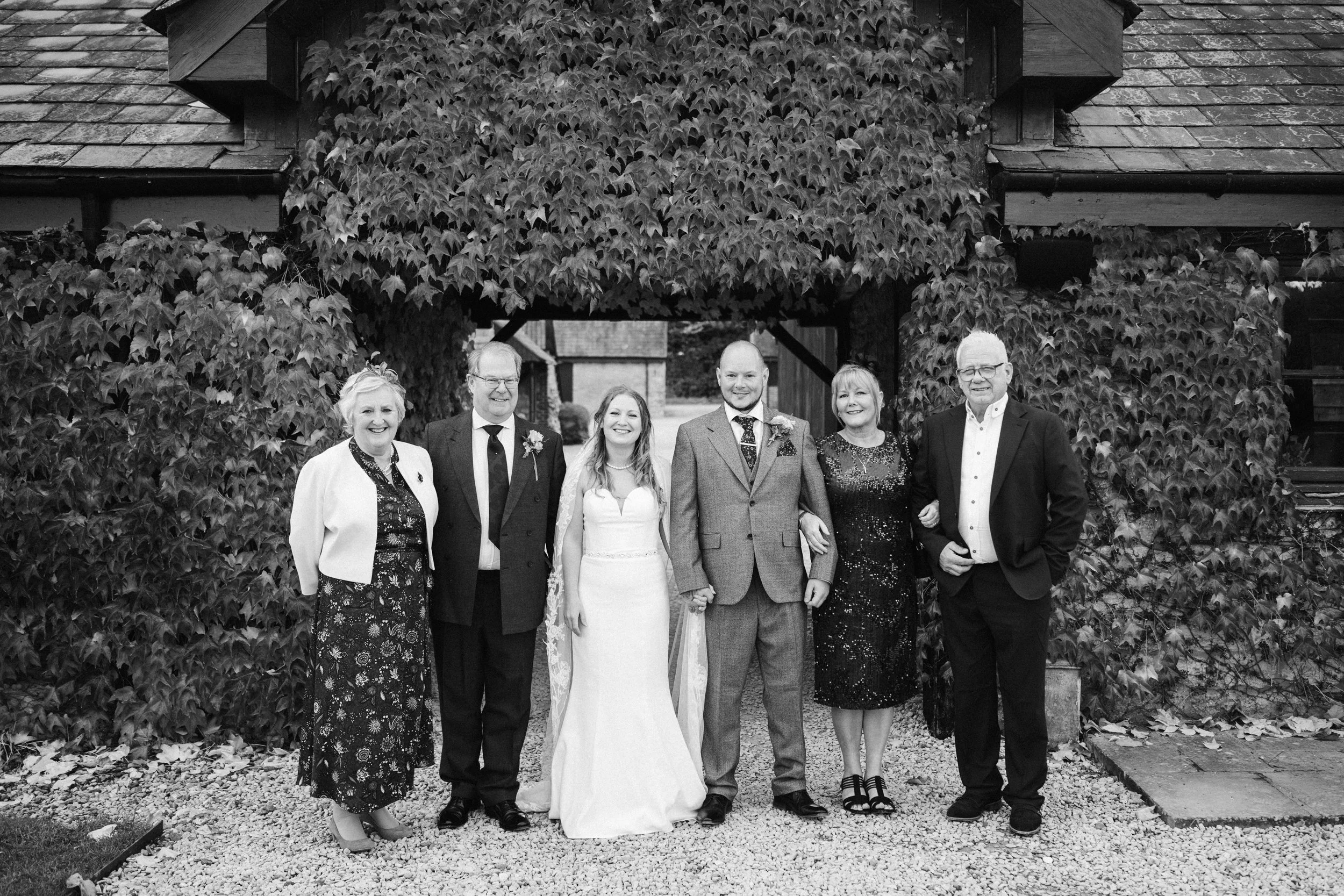 A family group photo outside in front of a leafy archway, with a bride and groom in the center. The bride is wearing a white wedding dress and the groom is in a gray suit. They are holding hands. Four other people in formal attire are standing beside