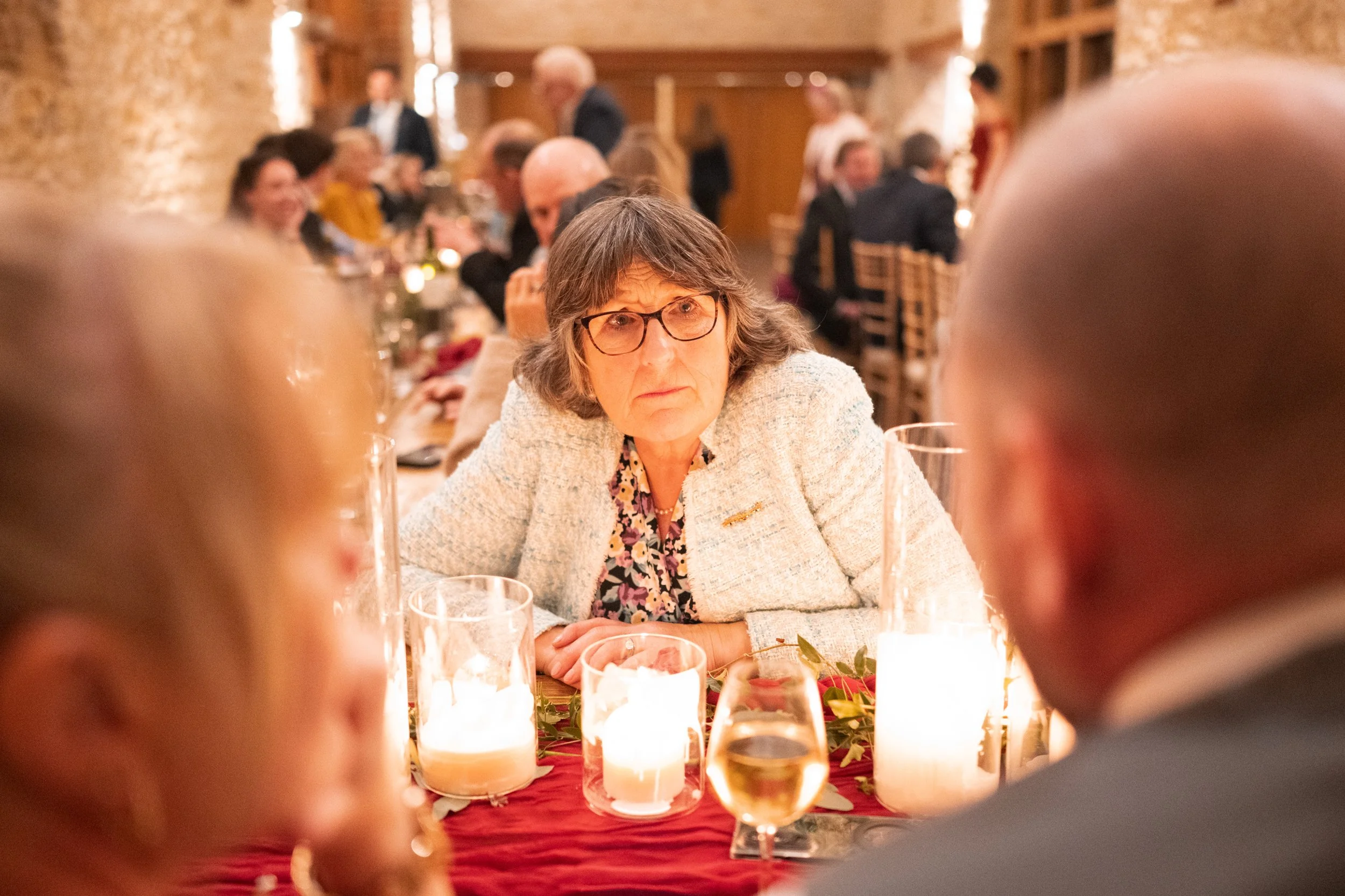A woman with glasses and gray hair looks concerned while sitting at a table with lit candles during a formal gathering or dinner event.
